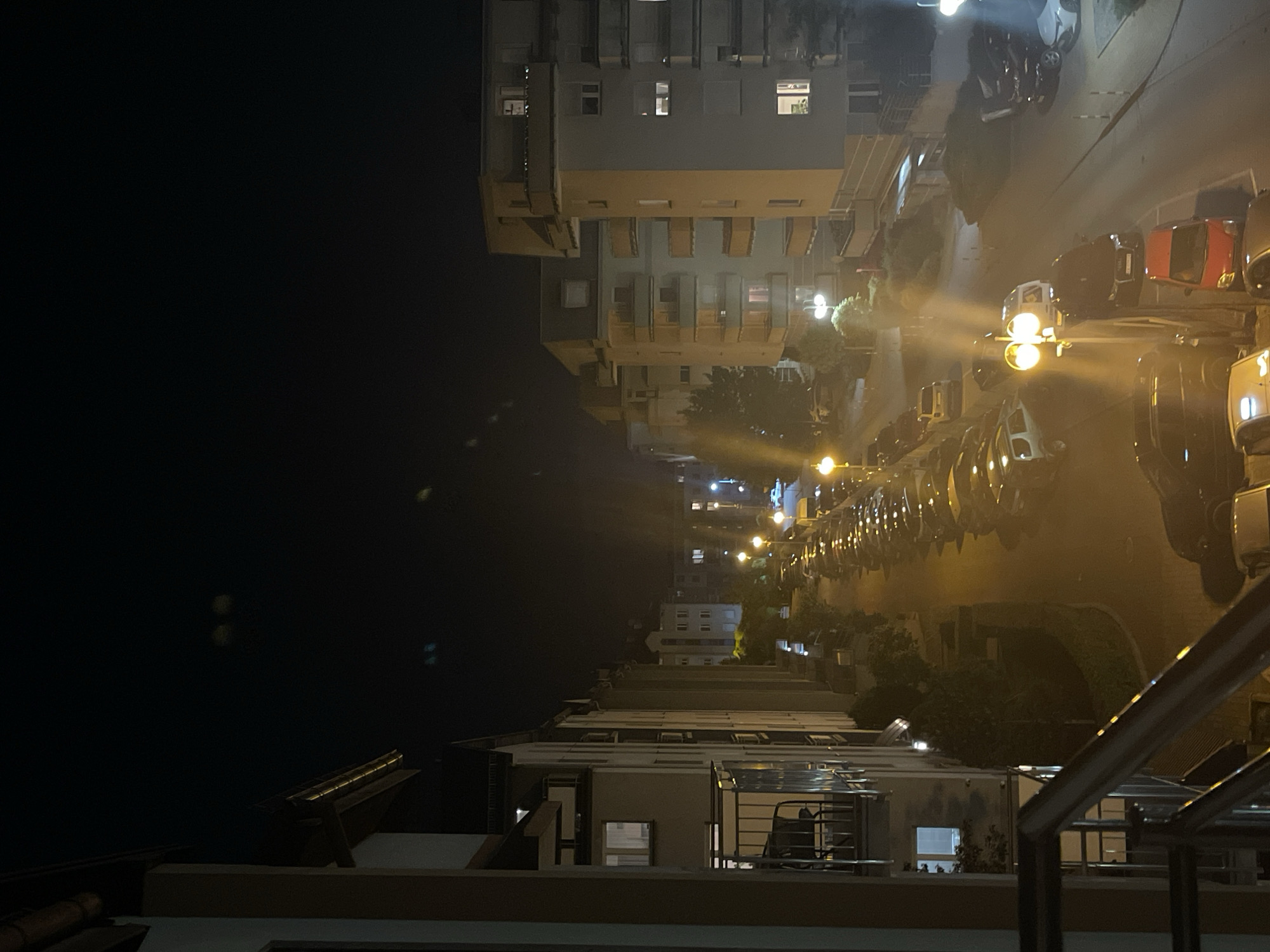 Here is a description of the image:
The image is an outdoor nighttime shot, showing an urban scene in Unknown, Unknown. In the upper right and middle right, a street is lined with parked cars and illuminated by streetlights. On the upper right and center is a multi-story building with lighted windows. In the upper left and middle left is dark water or sky. The bottom of the image shows part of a balcony with railings and architecture.