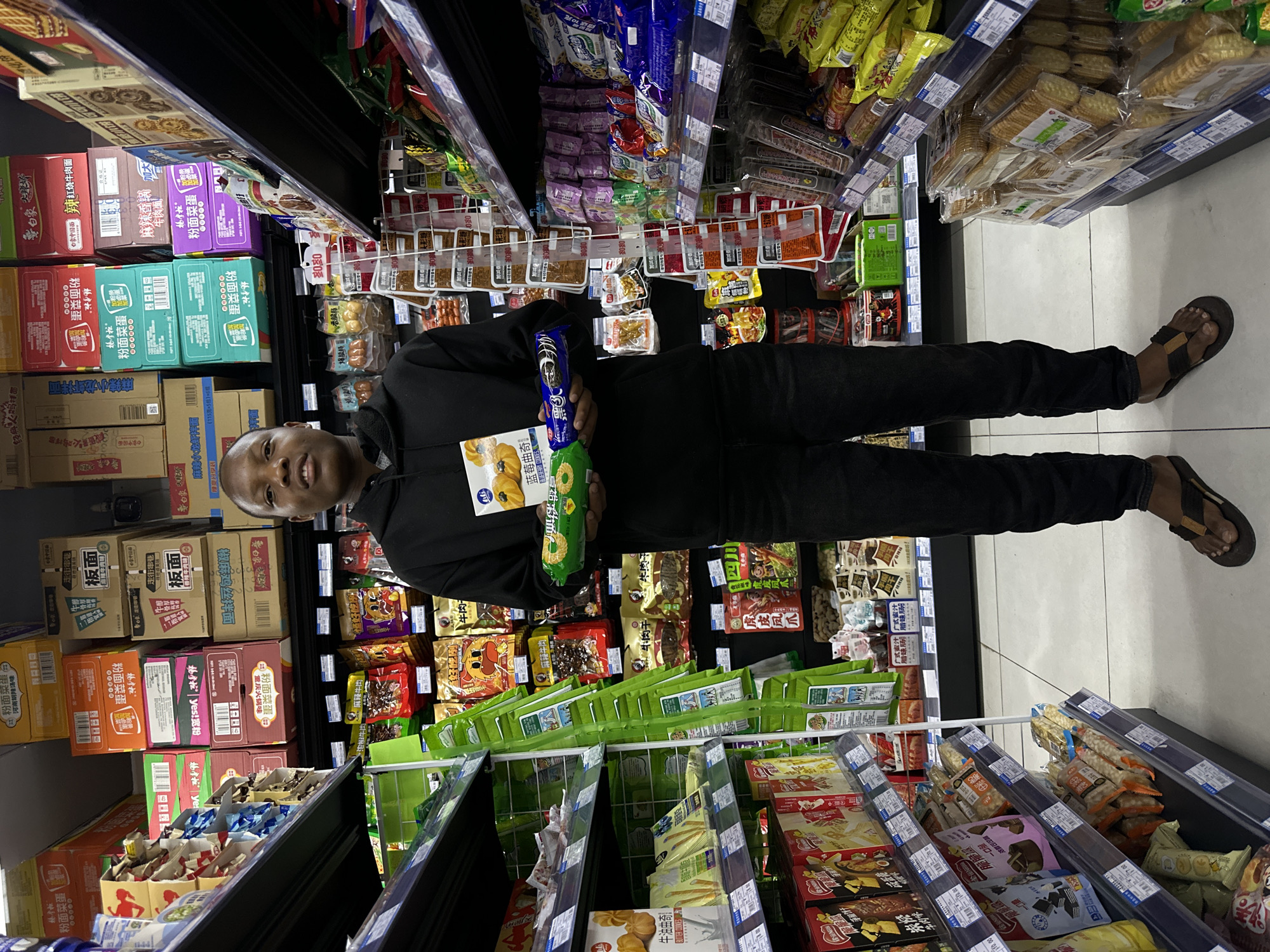 A person is standing in the aisle of a grocery store in Jiangjin, China. The person is wearing a black hoodie and black pants, and sandals. They are holding a blue package and a green package of snacks. The shelves are stocked with a variety of packaged food items.