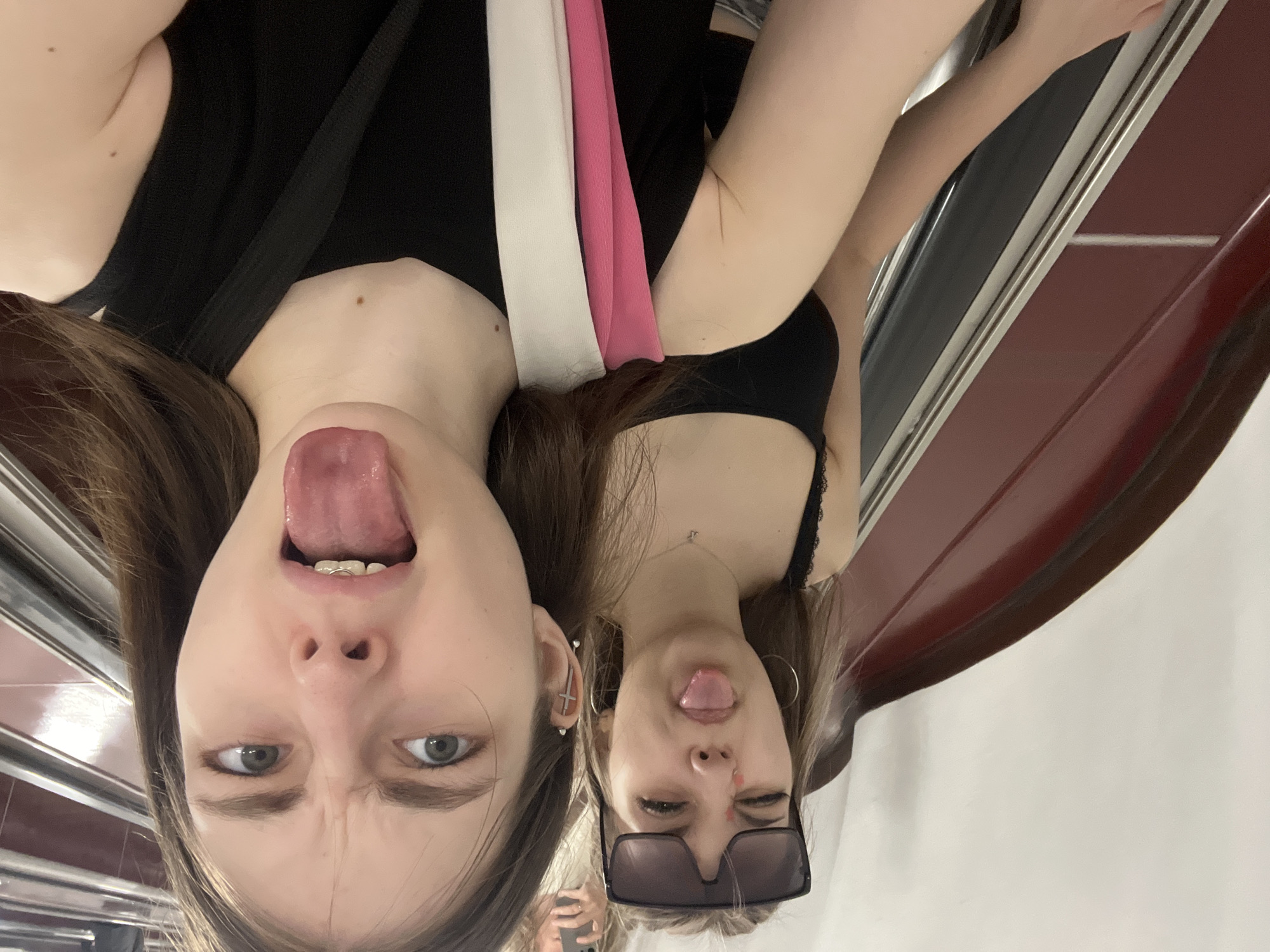Two young women are photographed upside down, likely in a public transportation setting. The woman on the left is sticking her tongue out and pulling a face, revealing braces on her lower teeth. She has a cross-shaped earring in her left ear and is wearing a black top with white and pink straps. The woman on the right is also sticking her tongue out, winking her left eye, and wearing sunglasses. She is wearing a black top with lace trim and a necklace. The background appears to be a train car interior, with a window and dark red paneling visible. The location is likely in Saint Petersburg, Russia, based on the visual cues present in the image.
