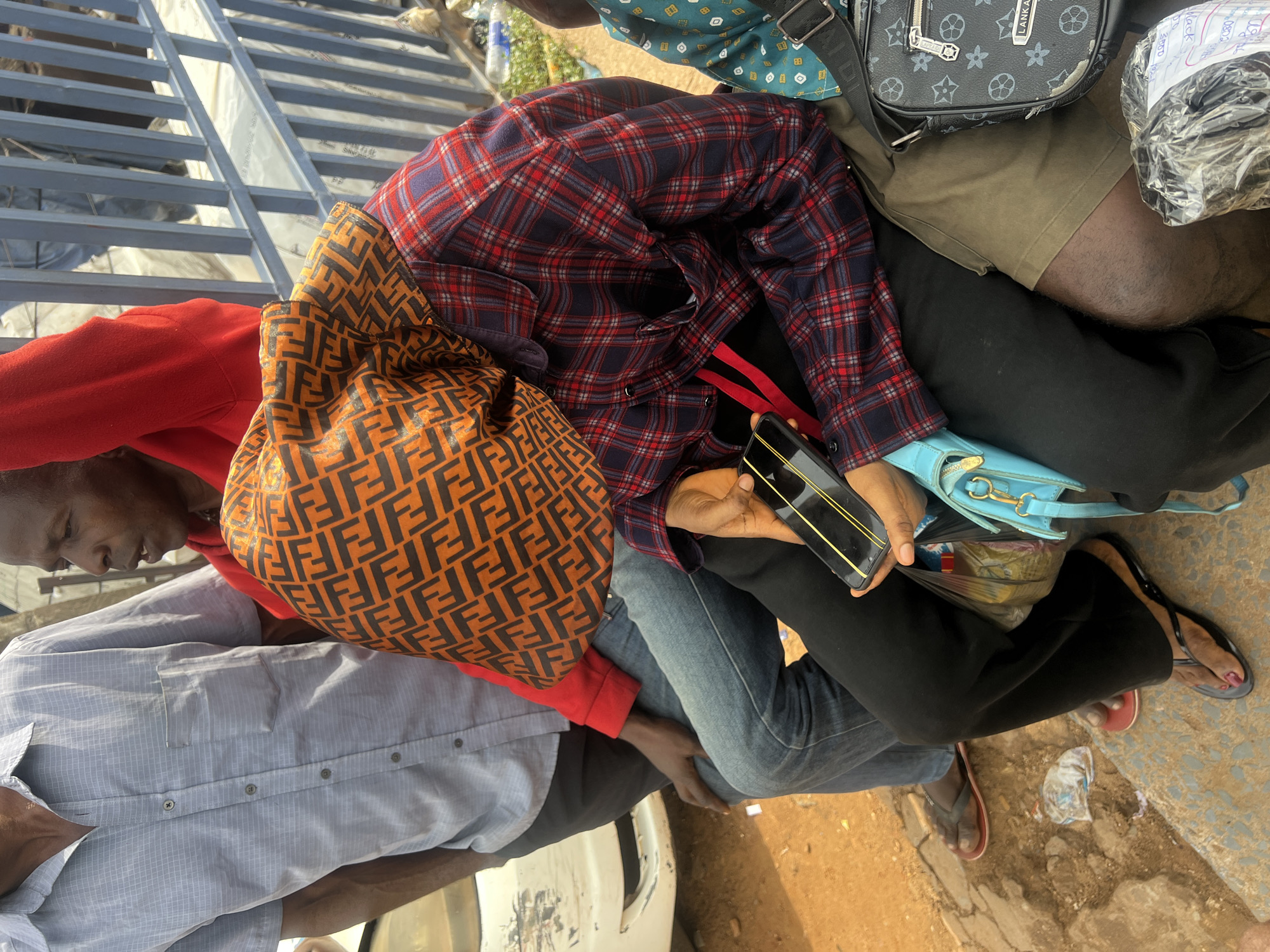 In Onitsha, Nigeria, three individuals are seated on a ledge. The person on the left wears a light blue button-down shirt and dark pants. Next to them, a person in a red hooded garment is mostly obscured by a patterned orange and black headscarf. This person is also wearing a red and navy plaid shirt and black pants, and is holding a smartphone. To the right, a person wearing khaki shorts is partially visible, with a black crossbody bag on their lap. The ground is covered in dirt and scattered debris. A white vehicle is parked to the left of the frame, and a blue metal railing runs along the background.