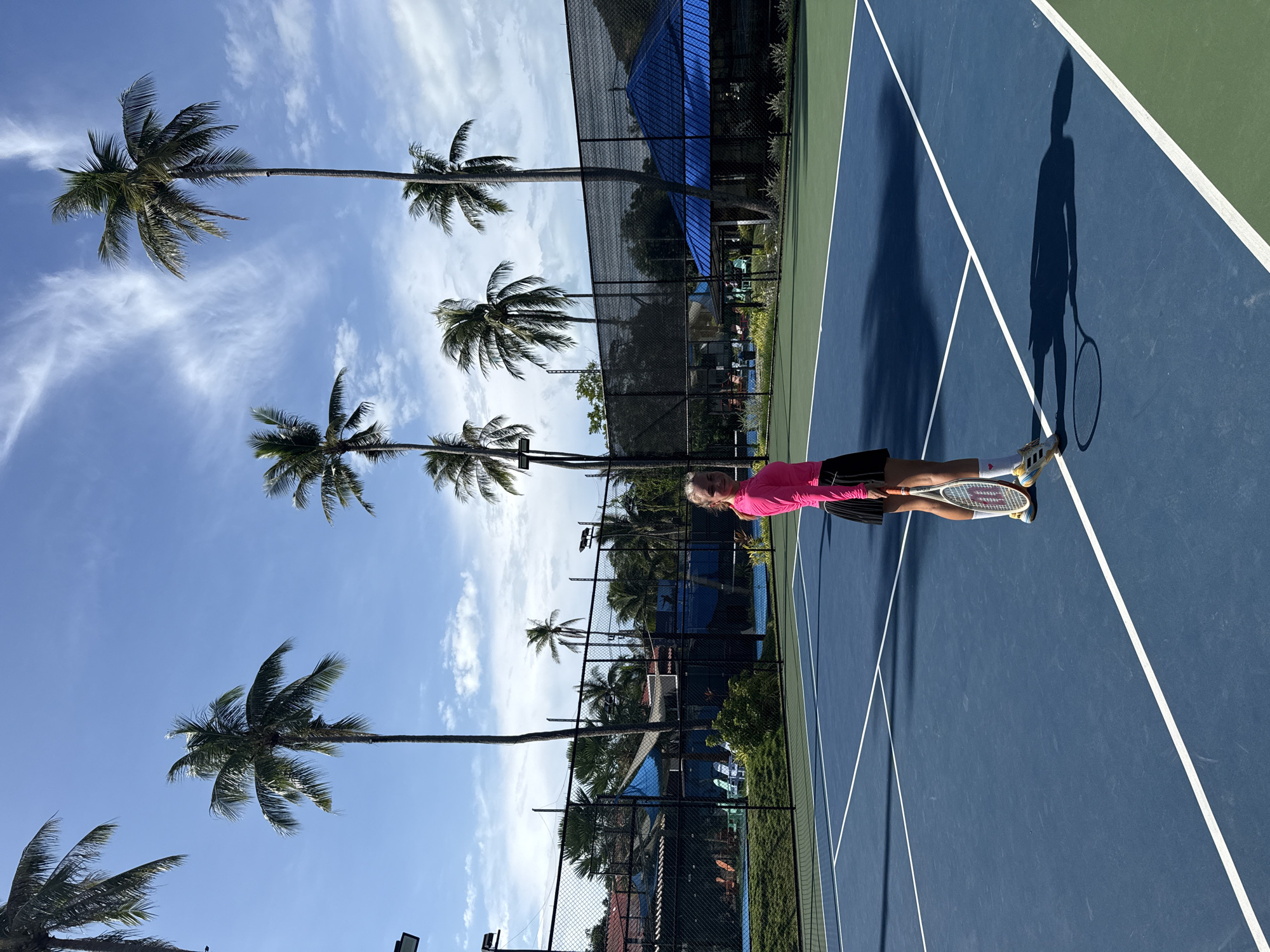 A young woman with blonde hair stands on a blue tennis court in Baan Plai Laem, Thailand, holding a tennis racket. She is wearing a bright pink long-sleeved shirt and a black skirt, with white socks and yellow tennis shoes. The court has white lines marking its boundaries. In the background, a black fence surrounds the court, beyond which are buildings with red roofs and a blue tent-like structure. Several tall palm trees reach towards a bright blue sky with scattered white clouds. The sun casts a long shadow of the woman and her racket onto the court. The environment suggests a tropical setting, likely a resort or sports facility.