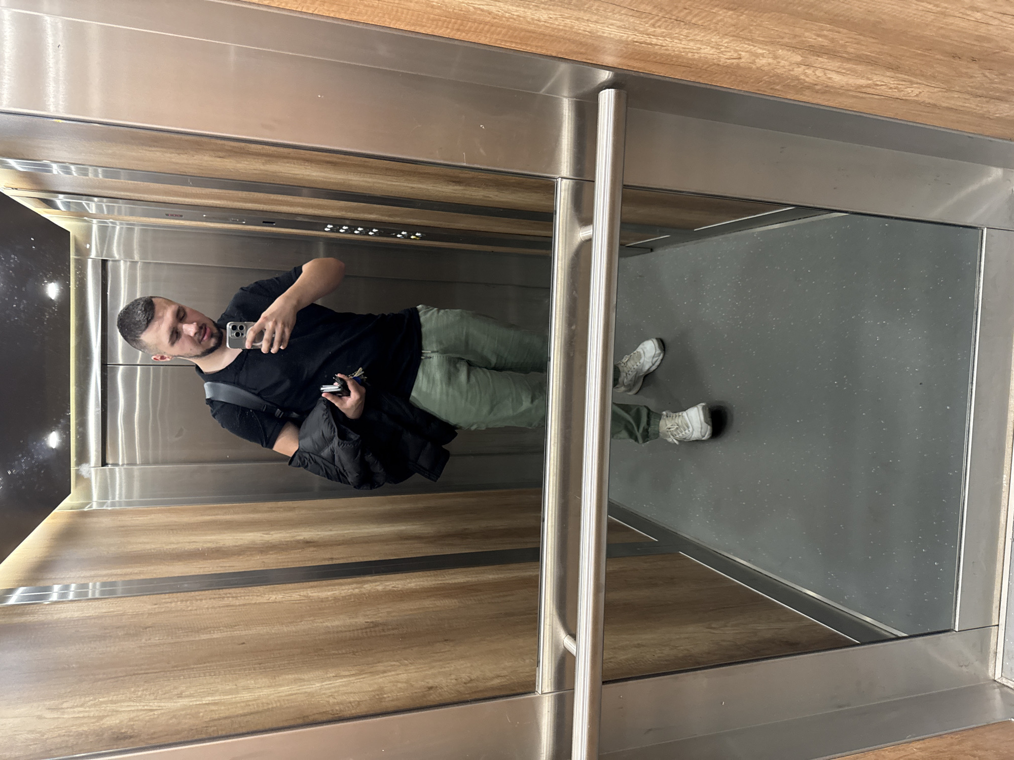 A young adult male with short dark hair and a beard is taking a selfie in an elevator. He is wearing a black t-shirt, green jogger pants, and white sneakers. He is holding a black jacket over his left arm and a set of keys in his right hand. The elevator has stainless steel walls and wooden accent panels. There are elevator buttons visible on the wall. The floor is dark grey. The image appears to be taken inside a building in Sofia, Bulgaria.