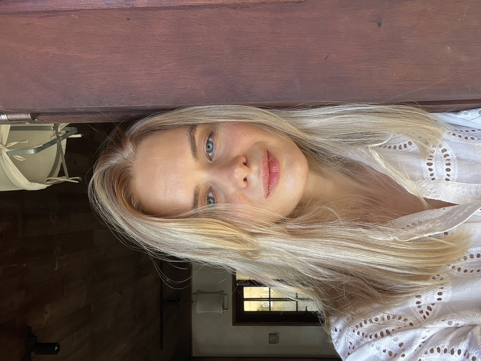 The image is a close-up selfie of a young Caucasian woman with long blonde hair and blue eyes. She is wearing a white embroidered shirt and leaning against a dark wooden doorframe. The background is dimly lit, showing a wooden ceiling and a doorway with a glass-paneled door and a sign. The woman's face is partially obscured by her hair, and she is looking directly at the camera with a slight smile. The scene appears to be indoors, possibly in a rustic or traditional setting. There are no other people or objects in the immediate vicinity that suggest a specific location within the city of Nha Trang Ward, Vietnam.