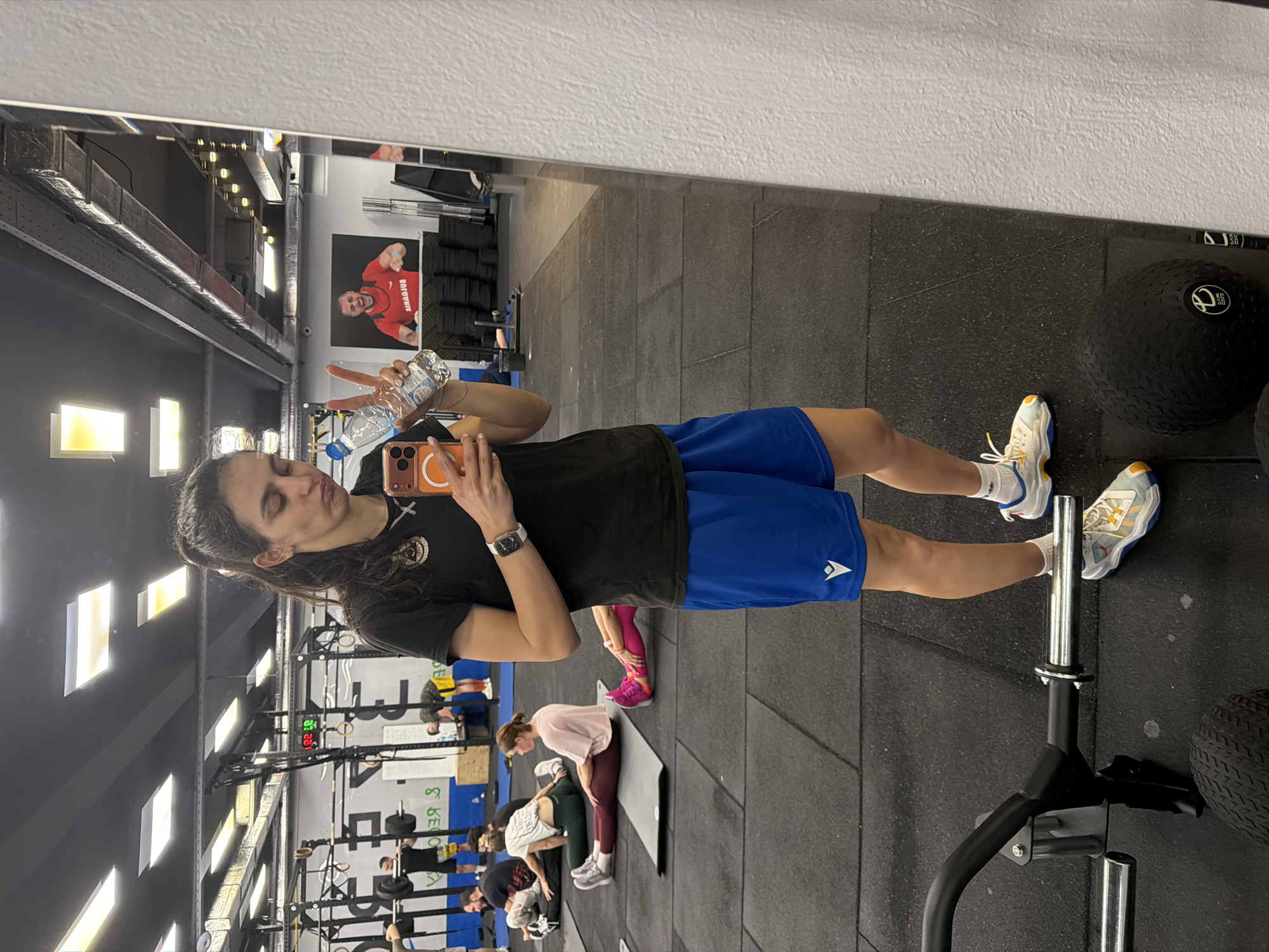A woman takes a selfie in a mirror in a gym. The woman has dark hair and is wearing a black t-shirt and blue shorts. She is holding a water bottle and making a peace sign. In the background, other people are working out. The gym appears to be located in Sofia, Bulgaria, given the "Airradius" advertisement featuring Bulgarian actor Stefan Popov on the wall. The gym features typical workout equipment like squat racks, barbells, and medicine balls.