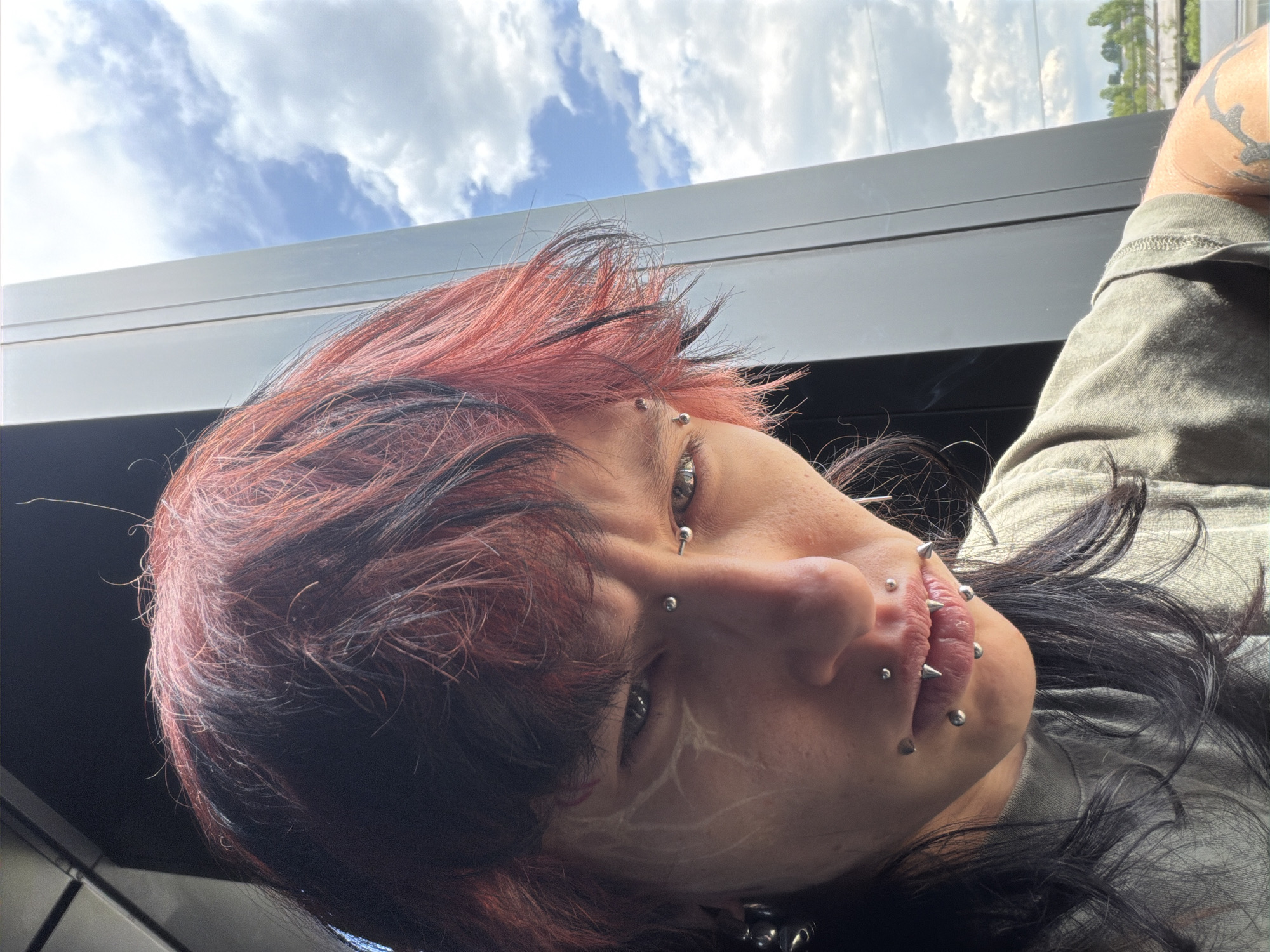 A close-up, slightly angled shot depicts a person with multiple piercings on their face, including around the lips and eyebrows. Their hair is styled with a combination of red and black strands. A white, vein-like design is visible on their cheek. The person appears to be positioned indoors, with a view of a cloudy sky and a glimpse of greenery visible through a window. The person is wearing a green shirt. The location of Busmantsi, Bulgaria, is not evident in the image.