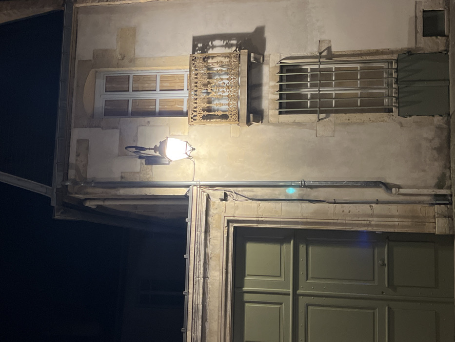 A building facade at night. A lit street lamp illuminates a gray stone wall with two windows on top, one with decorative metalwork, and a green wooden door below. A metal pipe runs horizontally above the door. The left side of the building is in shadow, contrasted with the light cast on the wall. Architectural details and textures are visible.
