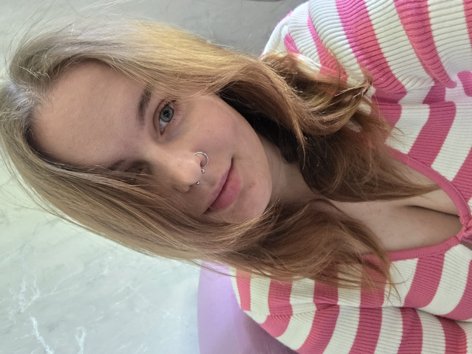 A light-skinned young woman with long, reddish-blonde hair, fair skin, and blue eyes, wearing a silver nose ring, is depicted in a close-up selfie. The woman is wearing a pink and white striped shirt. The environment appears to be a brightly lit indoor setting with a light-colored marble or tile surface visible. The city and specific location are unknown.