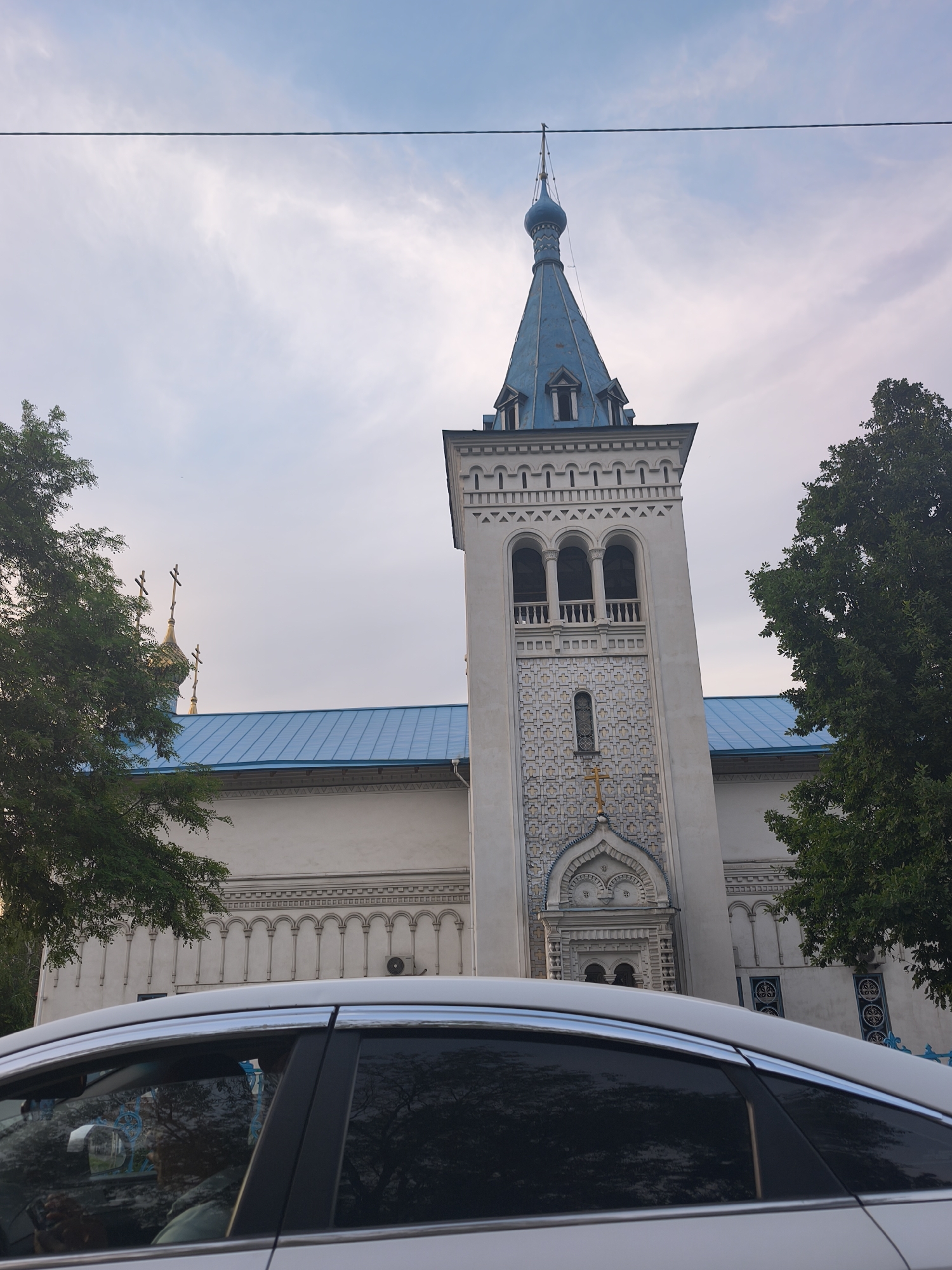The image presents an outdoor scene in Bishkek City, Kyrgyzstan, featuring an Eastern Orthodox church under a partly cloudy, soft blue sky. The time of day appears to be late afternoon or early evening due to the diffused lighting.
The central subject is a light-colored church building with a striking blue, possibly metal, roof. A prominent bell tower dominates the right side of the church, featuring arched openings for bells, intricate patterned tiling, and a small gold cross above an ornate entrance. The tower is topped with a blue, conical spire culminating in an onion dome and a cross. To the left, another section of the church's blue roof is visible, along with multiple golden onion domes, each topped with a cross.
Green trees with dense foliage partially frame the church on both sides, with a large tree on the right obscuring part of the building and another on the left providing a glimpse of the golden domes. A white or silver car is parked or driving in the foreground, occupying the lower third of the frame and partially obscuring the base of the church. A person is visible in the driver's seat of the car, holding a mobile phone. A dark electrical wire stretches horizontally across the top portion of the sky. No visible text is discernible in the image.
