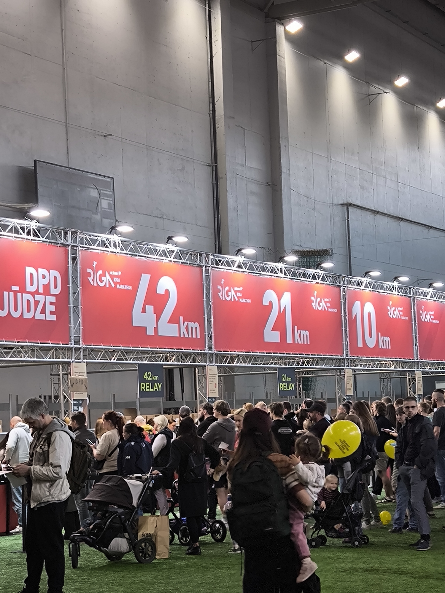 The image captures a scene within an indoor venue, potentially an arena or convention center, during the Riga Marathon in Riga, Latvia. The foreground shows a crowd of people, including adults with children and strollers, suggesting families are participating or supporting the event. Red banners suspended above the crowd display information about the marathon, specifying distances of 42 km, 21 km, and 10 km, each labeled with "Riga Marathon." Additional signage indicates relay races for the 42 km and 21 km distances. The artificial green turf underfoot simulates an outdoor environment. Overhead lighting illuminates the scene. The crowd is varied, with people engaged in different activities, from standing and talking to pushing strollers. The backdrop consists of a large, gray wall.