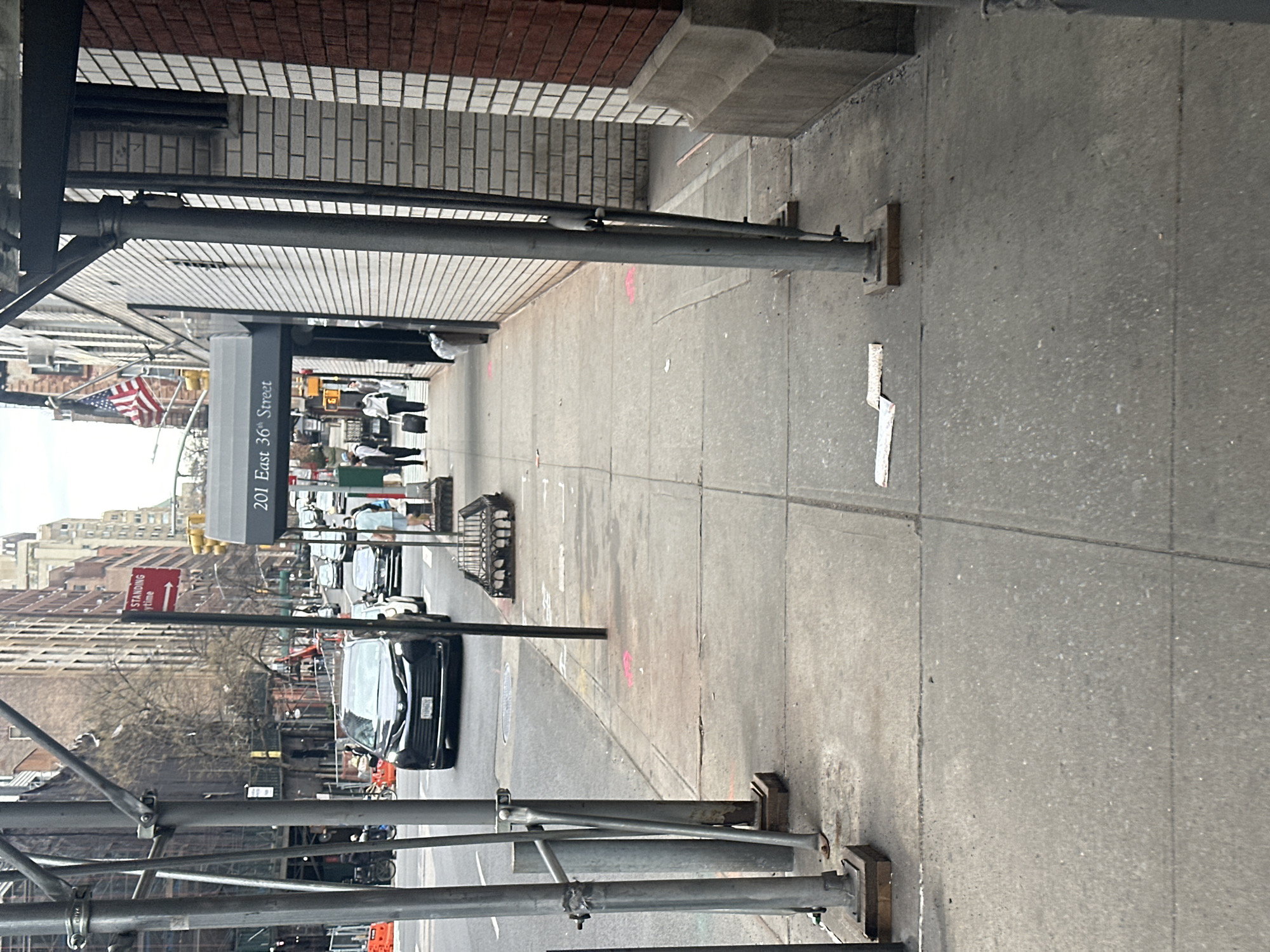This image depicts a daytime street scene on East 36th Street in New York City, United States of America, specifically at "201 East 36th Street." The perspective is from a concrete sidewalk, looking east down the street.
In the immediate foreground on the left, multiple grey metal scaffolding poles, secured with metal clamps and resting on wooden base blocks, extend vertically. The concrete sidewalk occupies the lower portion of the frame, showing minor surface wear and two pink spray-painted markings further down. On the right, a section of a light-colored brick building is partially obscured by scaffolding, with a reddish-brown brick building section visible beyond it. An American flag hangs from the light-colored brick building.
The street itself is visible beyond the foreground scaffolding. A black SUV, identified as a Toyota Sienna by its emblem, approaches the viewer in the left lane. Other vehicles are visible in the distance, receding down the street. A red "NO STANDING ANYTIME" sign is mounted on a pole near the street's left edge. An awning, prominently displaying "201 East 36th Street," extends from the light-colored brick building on the right.
Several individuals are present on the sidewalk near the awning. One person, wearing a light-colored top, pulls a black rolling suitcase. Another person in a light blue shirt is partially visible further down the sidewalk. A black metal grate, possibly for a tree pit, is visible on the sidewalk to the right of the red street sign. The overall environment suggests an urban setting with typical street furniture and ongoing activity.