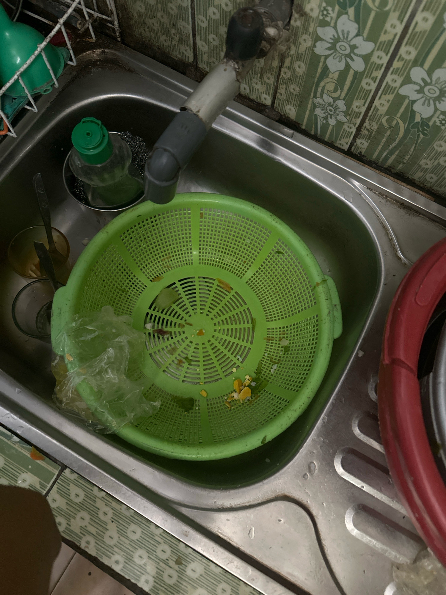 A top-down, high-angle shot shows a cluttered kitchen sink in Margamulya, Indonesia. The sink is filled with dirty dishes and food scraps. A green colander is overflowing with what appears to be vegetable scraps and some plastic packaging. To the left of the colander, there are two glasses, one partially filled with a yellow liquid and another empty. A bottle with a green cap and a metallic scrub brush are also present in the sink. A silver faucet is visible at the top of the sink, with a gray pipe connected to it. To the right of the colander, a red-rimmed pot or lid is partially visible. The sink is made of stainless steel and is stained with water marks. The backsplash behind the sink is tiled with a green and white floral pattern. The overall impression is one of disarray and neglect.