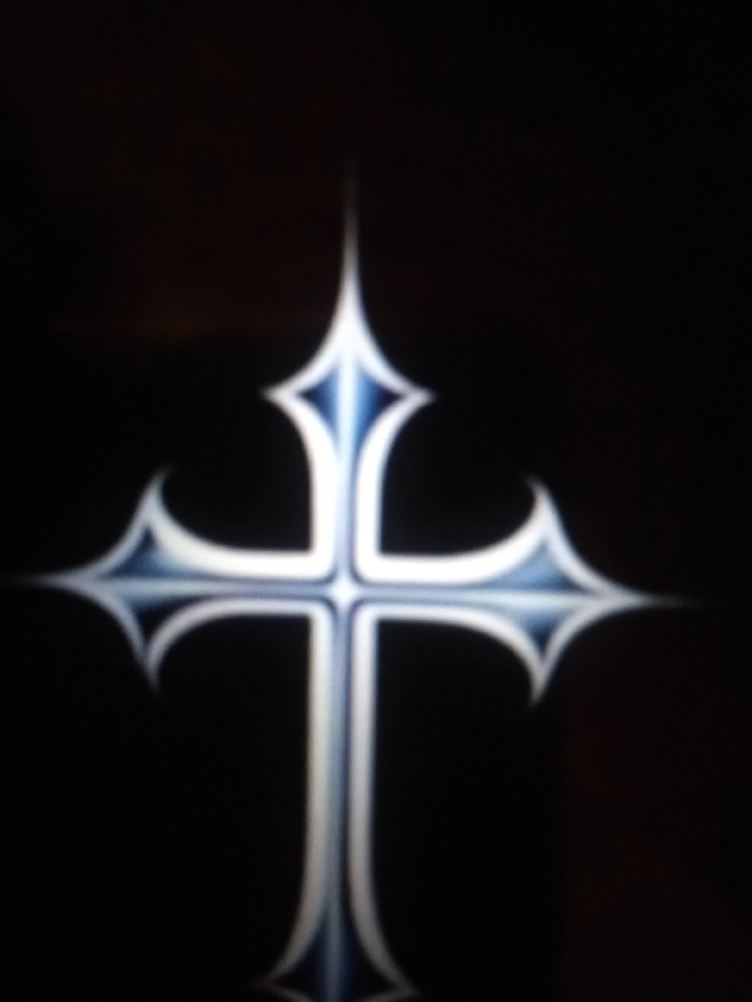 This media file is a close-up, blurred image of a stylized cross against a dark background. The cross appears to be illuminated with a blue and white gradient, giving it a luminous and somewhat ethereal quality. The shape of the cross is ornate, with pointed ends that flare outwards. The background is uniformly dark, offering no discernible environmental cues. There is no text visible in the image. The blurred nature of the image prevents any details about people, activities, time of day, weather, or specific location beyond the given context of Jalingo, Nigeria. The overall impression is one of a symbolic or decorative representation of a cross, presented in a visually striking but indistinct manner.
