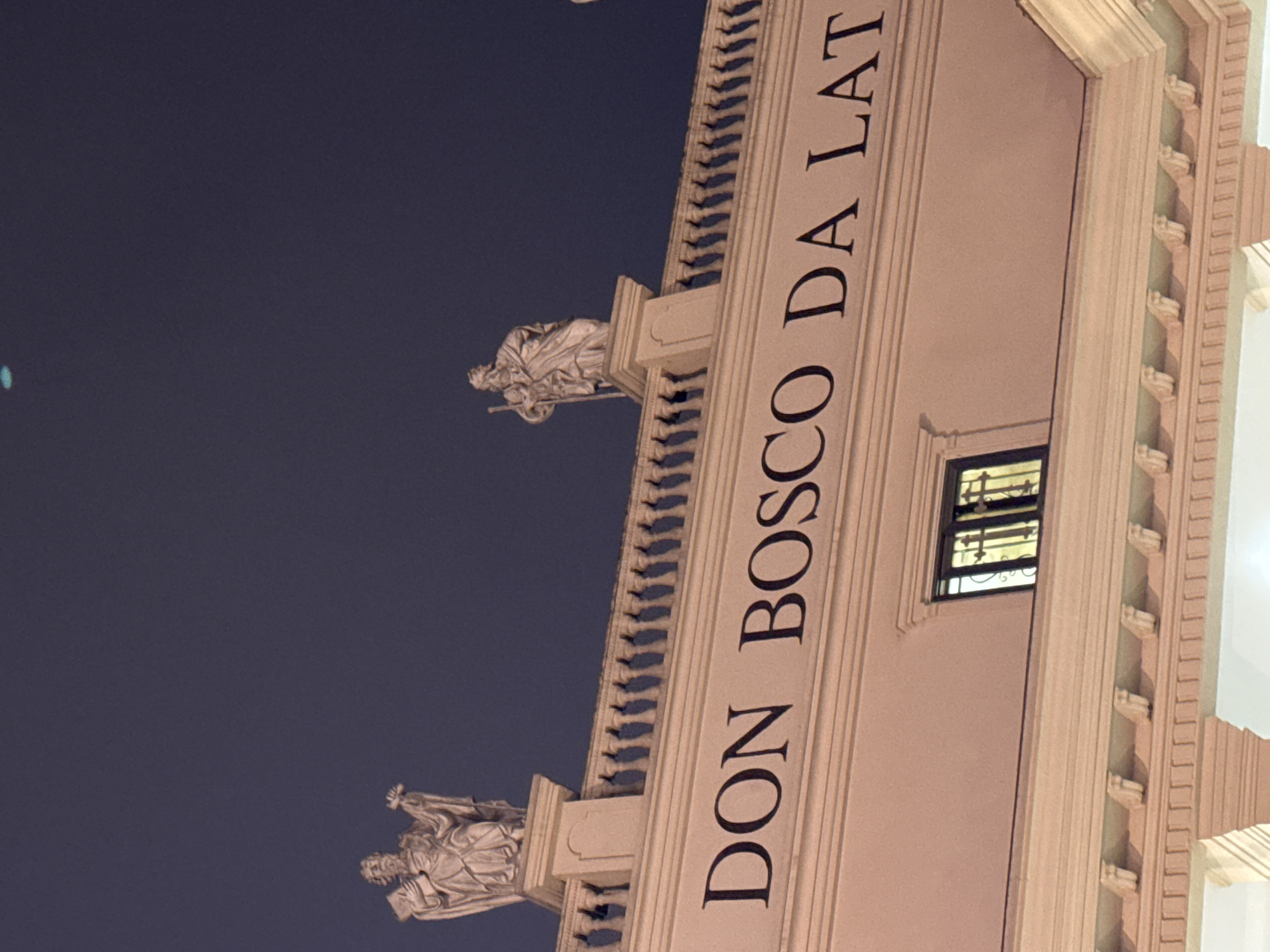 The image shows the facade of a building, "DON BOSCO DA LAT" inscribed vertically on the facade. Two statues are placed on ledges. A window is visible on the facade. The sky is dark. This appears to be a view of Don Bosco Da Lat Catholic Church located in Da Lat, Vietnam.