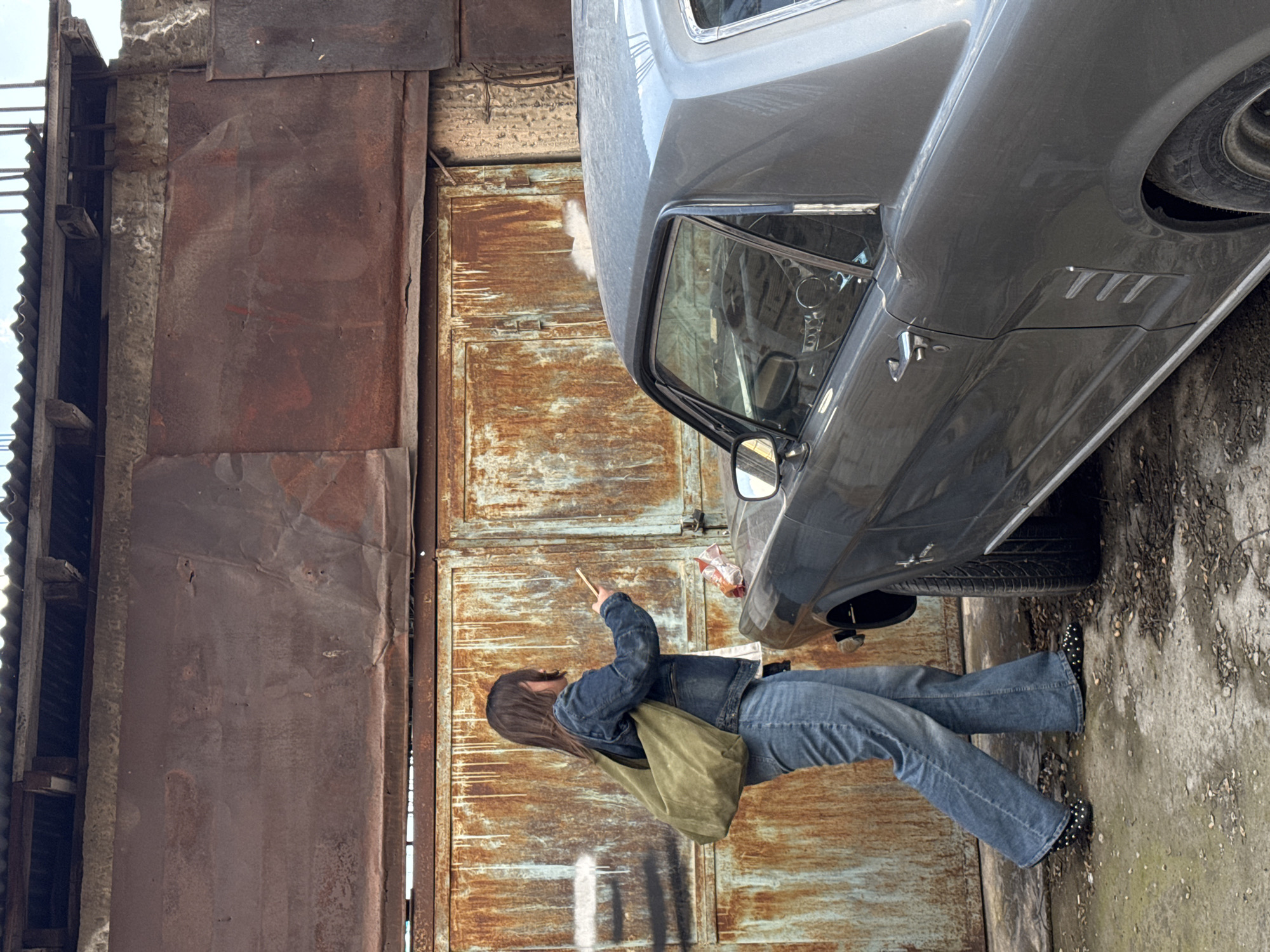 A medium shot depicts a woman with long, dark hair, wearing a denim jacket and jeans, standing on a concrete surface next to an old, gray car. She is holding a smartphone in her right hand, appearing to take a photo or video. A large, olive green canvas bag hangs on her shoulder.
The background consists of a heavily rusted metal wall with what looks like a rusty door. Part of a corrugated metal roof is visible at the top left of the frame. The image appears to be taken in an urban environment, possibly in Yerevan, Armenia given the user context.