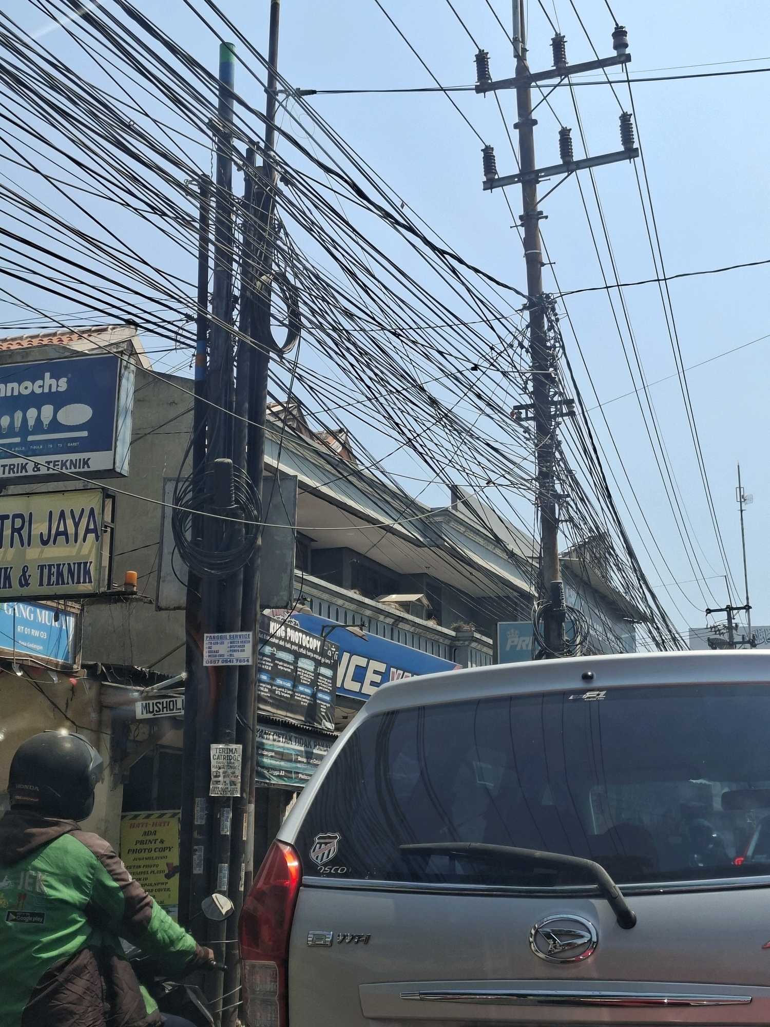 The image depicts a street scene in Calukan RT 1 RW 5 Keboansikep, Indonesia, characterized by a dense network of overhead electrical and telecommunication wires. A silver car is visible in the foreground, occupying the right side of the frame, facing away from the camera. The rear windshield of the car displays some reflections and a wiper. To the left of the car, a person wearing a green jacket and a black helmet is seated on a motorcycle, facing away from the camera. Several buildings are visible in the background, with some featuring signage. One sign, on the left, reads "nnochs" and appears to be an advertisement for electrical products. Another sign, in yellow, says "TRI JAYA." A blue sign with white text is also present. The scene appears to be set during the daytime under a clear sky.