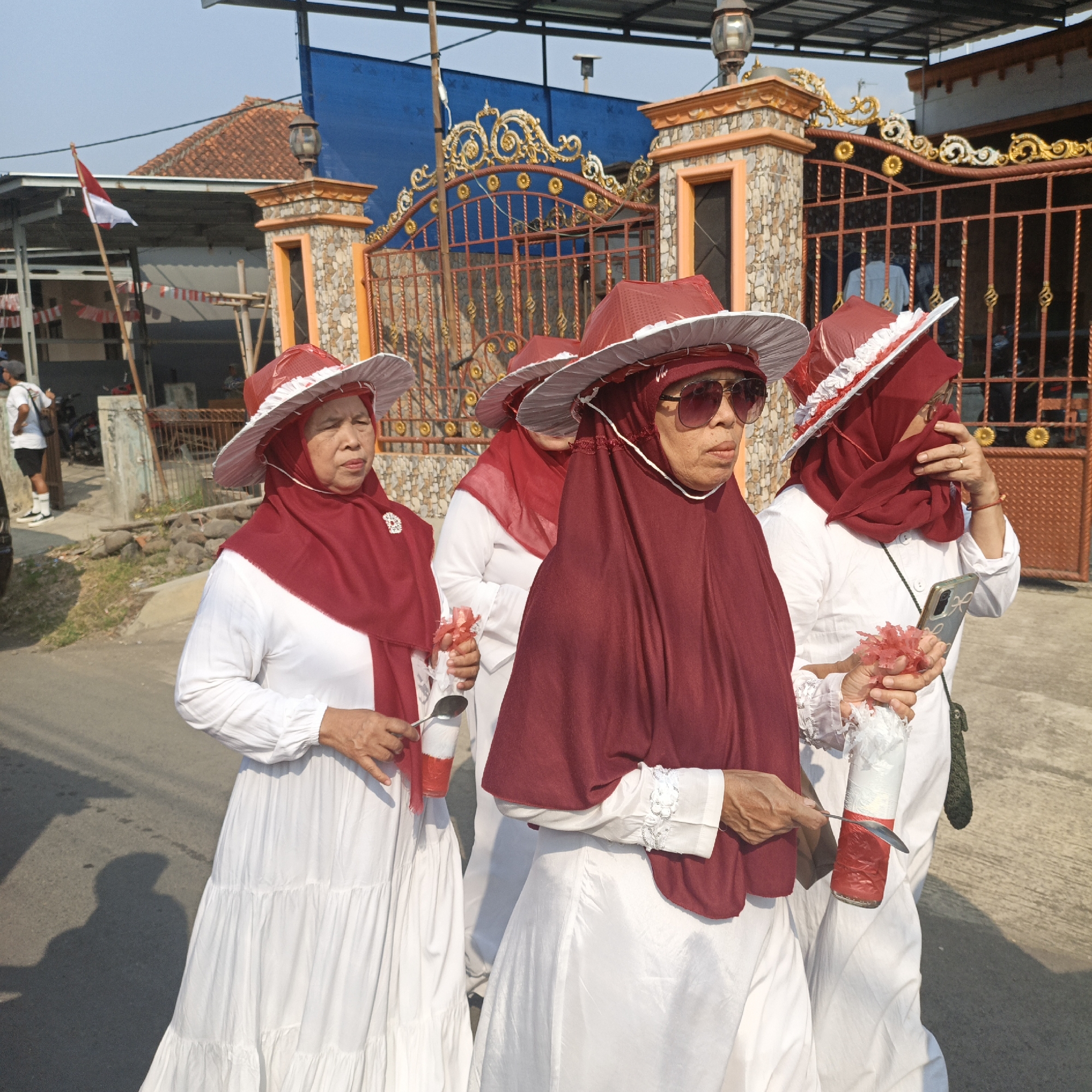 In Tanahbaya, Indonesia, a group of four women are walking along a paved street. The women are all wearing white dresses and red hijabs, and three of them are wearing red and white wide-brimmed hats. The women appear to be participating in a parade or celebration, as evidenced by the Indonesian flag flying in the background and the festive decorations on a nearby gate. The woman in the foreground is wearing sunglasses and holding a cell phone. The other women are holding small decorative items. The environment is a residential area with houses and a gate visible in the background. The sun is shining, casting shadows on the street.
