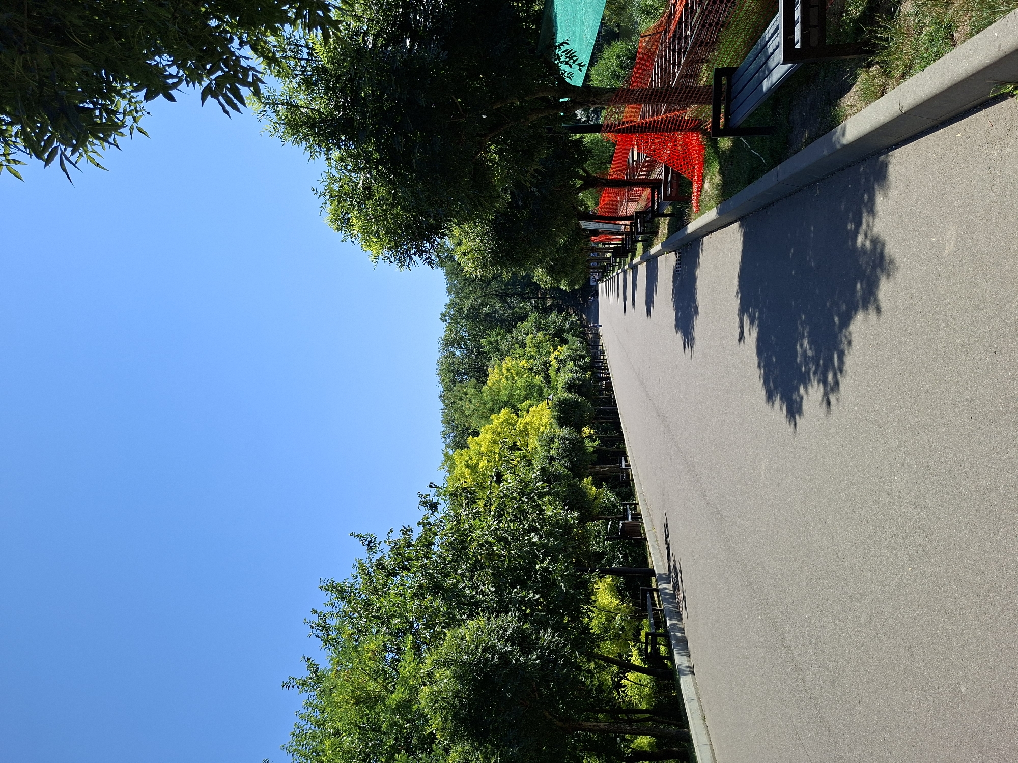 The image shows a paved pathway in a park on a sunny day. The pathway curves to the right and leads uphill. On the left side of the pathway, there are trees with green foliage and some benches. On the right side, there are more trees, benches, and an area with orange safety fencing and a green tarp. There are a few people visible in the distance on the pathway. The sky is clear and blue. The location is not specified in the image.