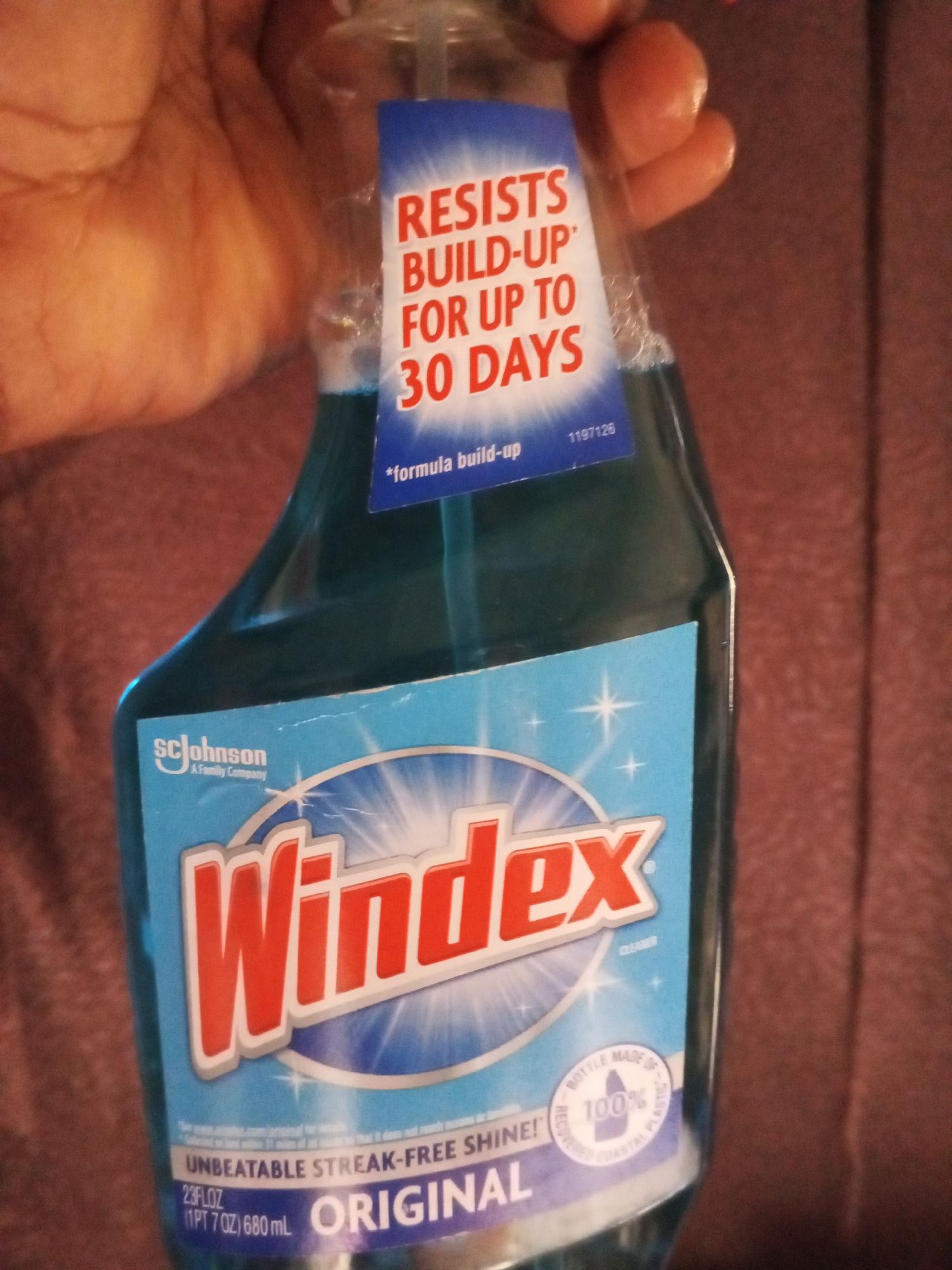 An image shows a Windex bottle held in a person's hand. The bottle is blue and transparent, with a blue label that has white and red text. The text on the label includes "Windex," "Resists Build-Up For Up to 30 Days," and "Unbeatable Streak-Free Shine." A small white circle on the label indicates "Bottle Made of 100% Recycled Plastic." The background is a blurred reddish-brown surface, possibly upholstery. The image does not contain any information about the city of Ashata, Nigeria.