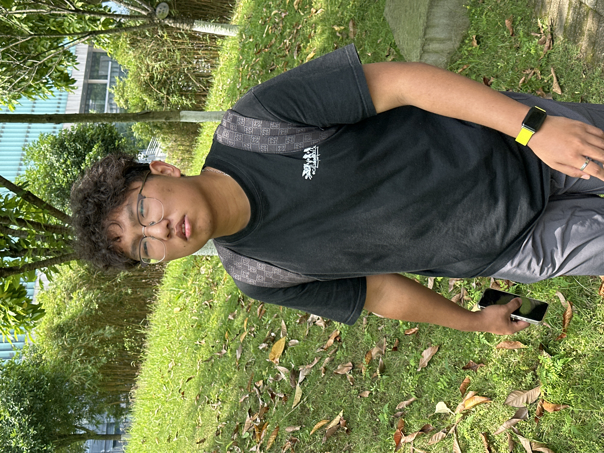 A person stands in a grassy area with scattered fallen leaves. The person is wearing a black t-shirt with a graphic on the front, grey shorts, and glasses. They have a black watch with a yellow band on their left wrist and are holding a mobile phone in their right hand. The background consists of greenery, including trees and shrubs. A building is partially visible in the distance. The location is Jiangjin, China.