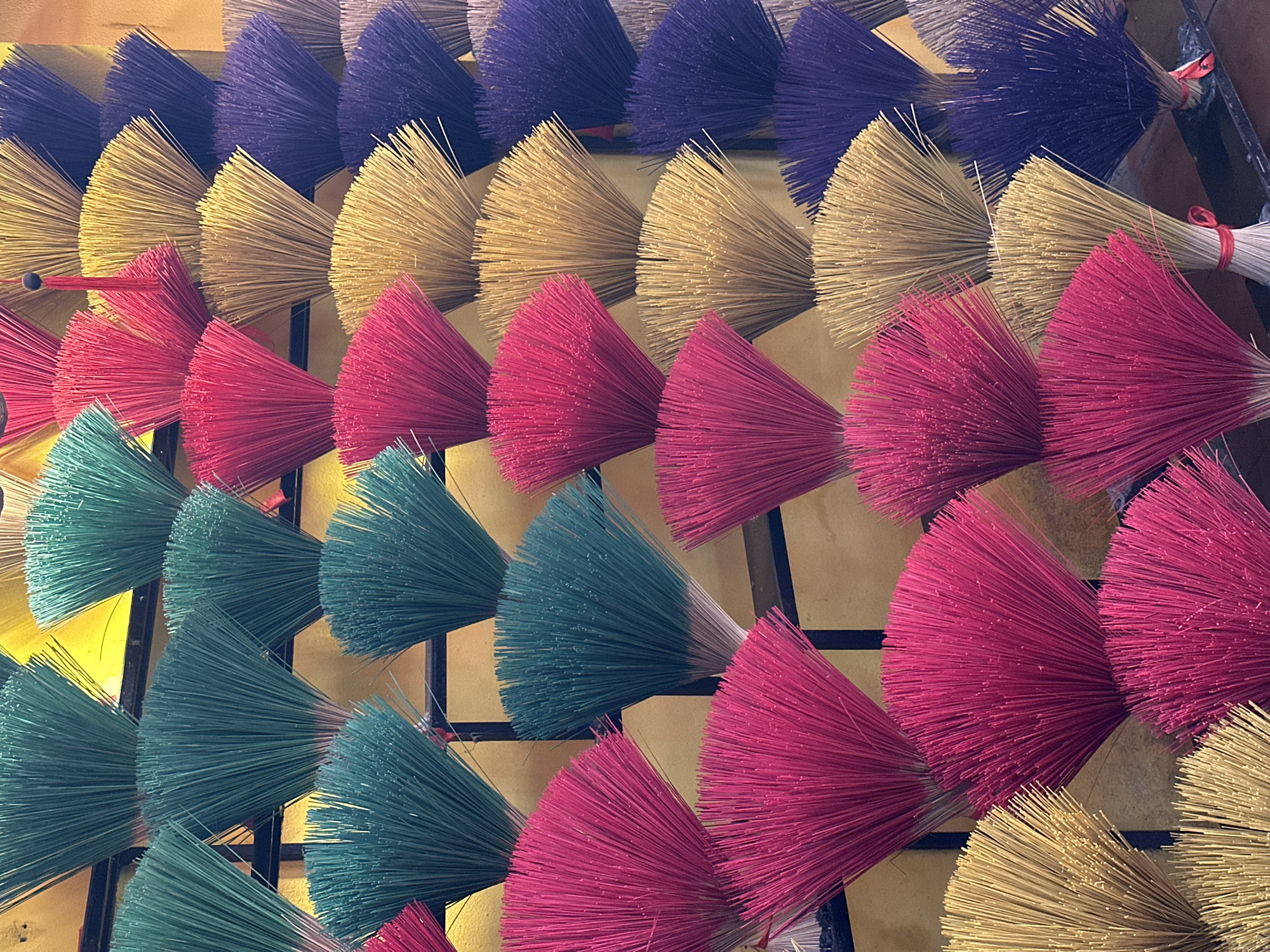 A high-angle, close-up shot displays rows of colorful bundles, likely incense sticks, organized in a grid pattern. The bundles are arranged in distinct color rows: purple, gold, red, green, red, gold, red, and green. Each bundle is tied tightly at the base, creating a fan-like spray of fibers. The backdrop is a muted yellow, possibly a wall or display surface. Black grid lines are visible behind the bundles. The setting suggests a vendor's stall, with the arrangement aimed at catching the eye. Without additional context, specifying the location in Đà Nẵng, Vietnam, is not possible from the image alone.
