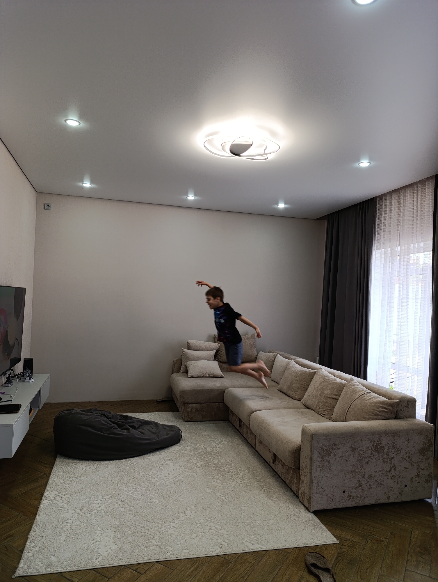 This medium eye-level shot captures a modern living room, likely in Chaikovsky, Russia, during the daytime with artificial lights also on. The room features light-colored walls and dark parquet flooring, partially covered by a large, light-toned patterned rug.
A young boy, dressed in a dark t-shirt and shorts, is frozen in mid-air as he jumps playfully over a large, light-beige L-shaped sectional sofa. His arms are slightly raised, and legs bent, conveying energetic motion. The sofa has multiple plump cushions.
To the left, a flat-screen television sits on a low, white media console. A dark gray beanbag chair rests on the rug between the television area and the sofa. The ceiling is equipped with multiple recessed spotlights and a decorative central light fixture, all illuminated.
On the right side of the room, a large window is adorned with a combination of dark gray opaque curtains pulled to the sides and a sheer white curtain that diffuses the incoming daylight. A single dark slipper is visible on the wooden floor in the bottom right corner. The room appears tidy and well-lit, designed for comfort and activity. No visible text is discernable.