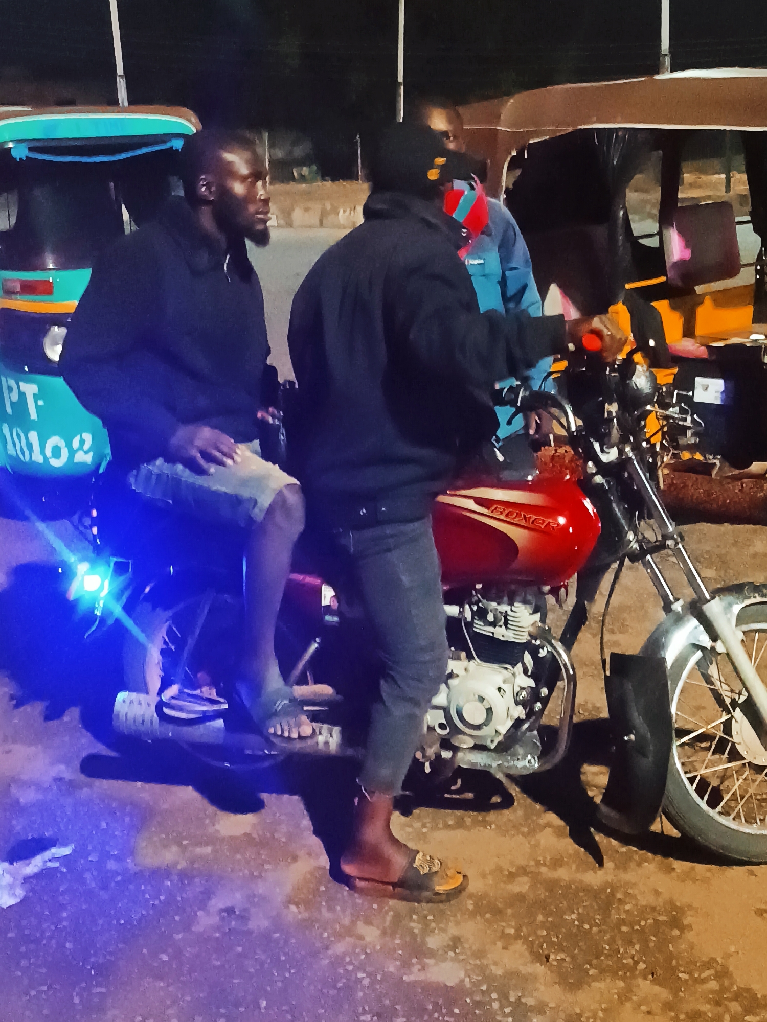 Three men are on or around a red Boxer motorcycle in what appears to be a street in Bukuru, Nigeria, at night. One man sits on the motorcycle, wearing dark clothing and flip-flops. Another man, also in dark clothing, stands next to the motorcycle. A third man stands between them wearing a blue jacket. The motorcycle is positioned between two tuk-tuks, one with the registration "PT-18102". The background suggests an urban or semi-urban environment with street lighting.