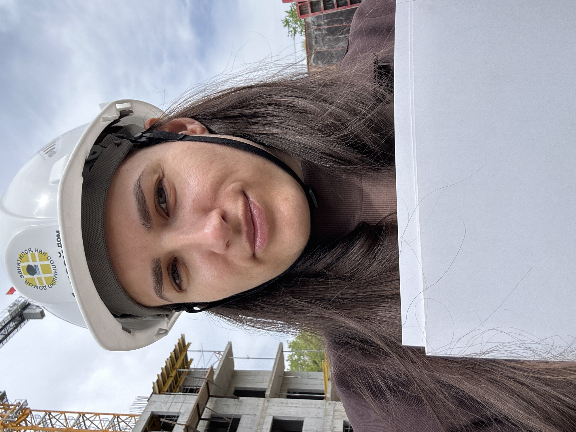 A woman wearing a white hard hat is posing for a selfie in front of a construction site. The hard hat has text in Russian: "закатилось, как солнышко домой". In the background, construction of a building is visible with a yellow crane. Based on the text on the hard hat and construction style, the location is in the city of Khabarovsk, Russia. The woman is holding white sheets of paper.
