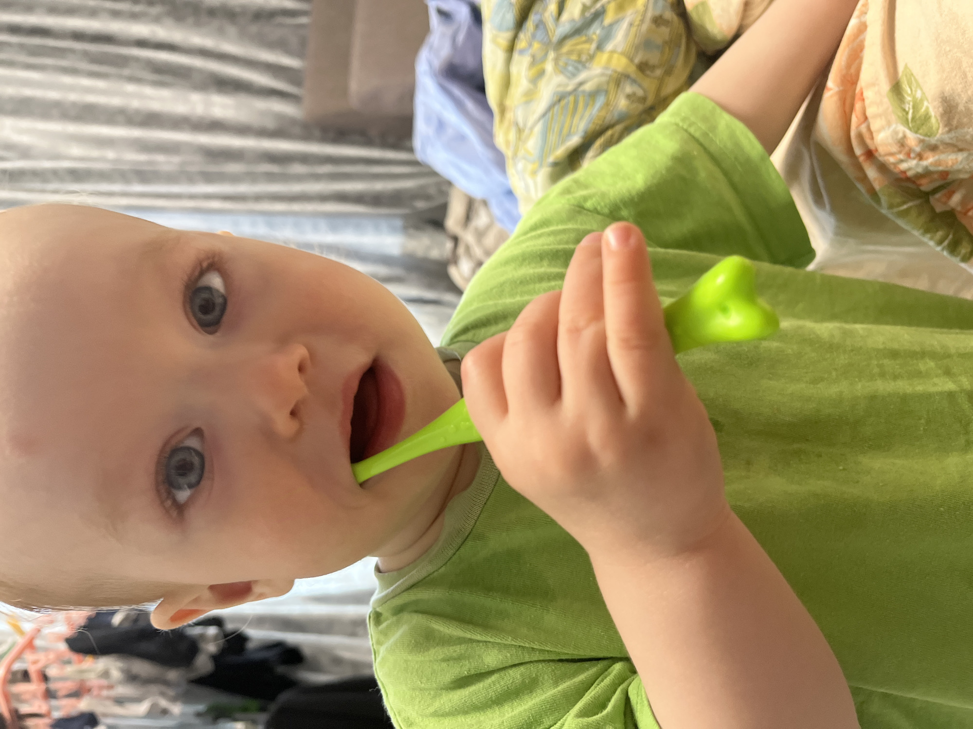 A young child, seen from a close-up, high-angle perspective, is holding a green object in their mouth. The child has light skin, blue eyes, and fair hair. They are wearing a green shirt. The background includes bedding with a gray and white striped pattern, as well as other linens with various colors and patterns. There is no information about the location being the city of Chaikovsky, Russia in the image.