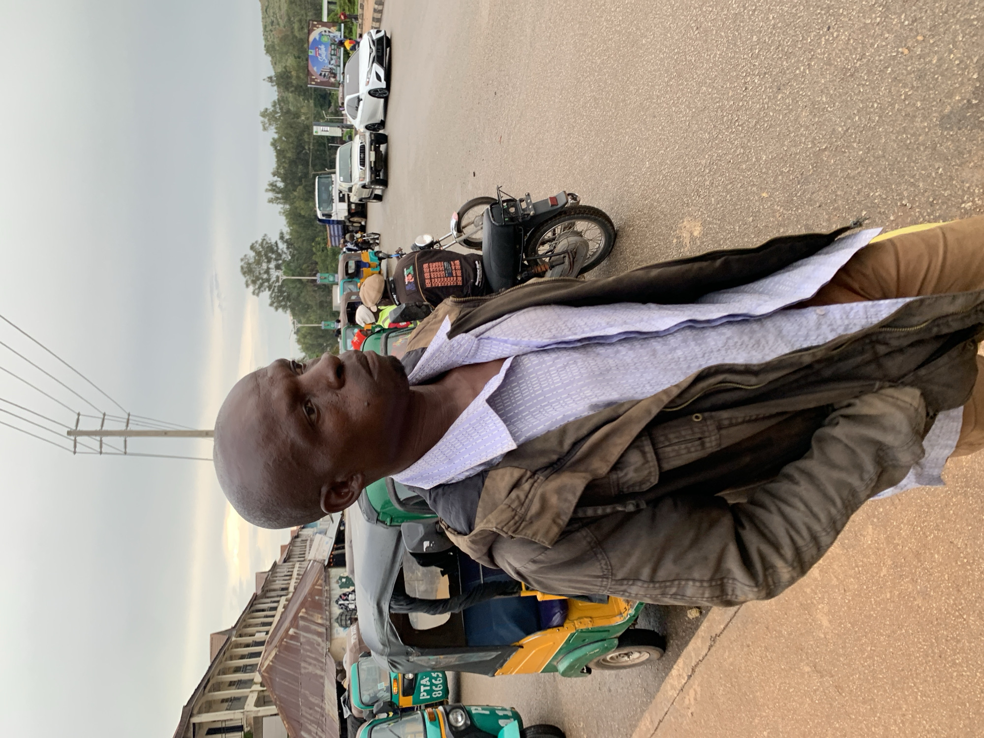 The image captures a street scene in Gura Top, Nigeria. In the foreground, a man with a shaved head is standing and looking to his right, wearing a dark jacket over a light blue patterned shirt and tan pants. Behind him, several auto-rickshaws are parked or moving, one of which is yellow and green with "PTA 8665" visible. Further in the background, other vehicles, including cars and a truck, are on the road. Buildings with multiple stories line the left side of the street, featuring weathered facades and awnings. The sky is overcast, and the road surface appears to be asphalt.