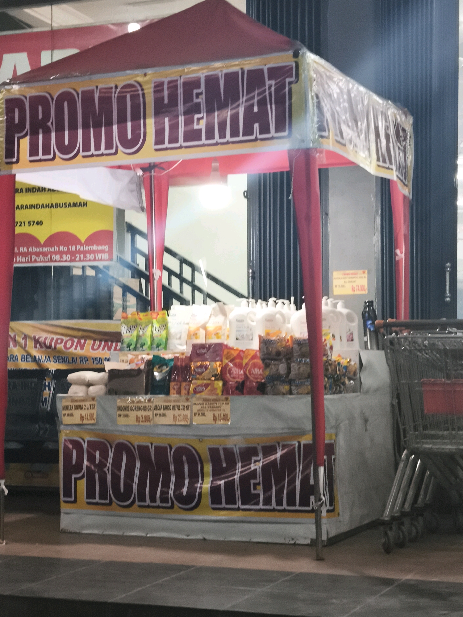 The image shows a promotional stall set up outside a store, likely in Palembang, Indonesia, given the text in Indonesian. The stall is covered by a red and yellow tent with a banner that prominently displays "PROMO HEMAT" (Discount Promotion). Various products are displayed on shelves, including what appear to be bottles of liquid soap or detergent, packaged snacks, and cooking oil. Some of the products have price tags in Indonesian Rupiah (Rp). In the background, there are shopping carts and a closed storefront with vertical metal doors. The overall scene suggests a retail environment during a promotional event.