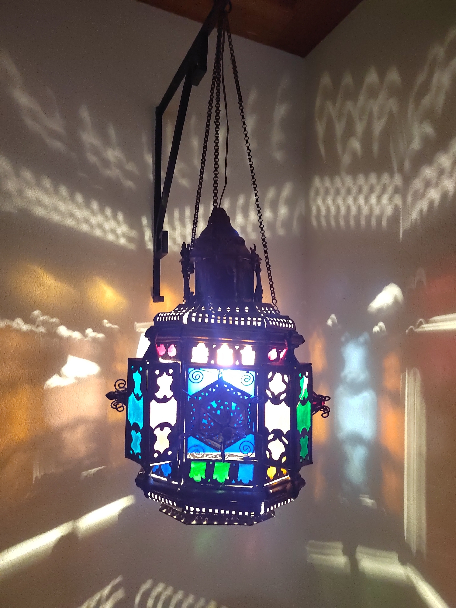 This is a close-up, eye-level, indoor shot of an ornate, multi-colored glass lantern hanging from a dark metal chain. The lantern is the central subject, its intricately designed metalwork and vibrant glass panels (including blue, green, and red) illuminated from within. The lantern casts dramatic, abstract shadows and colored light patterns onto a textured, light-colored wall and ceiling. The shadows appear in wavy, cloud-like, and geometric formations. The top of the image shows a glimpse of a wooden surface, possibly a ceiling beam, and a dark metal bracket from which the chain suspends the lantern. The overall atmosphere is one of warm, atmospheric lighting. The location of this scene is confirmed to be in the city of Porto Recanati, Italy. There are no people or animals visible in the image.