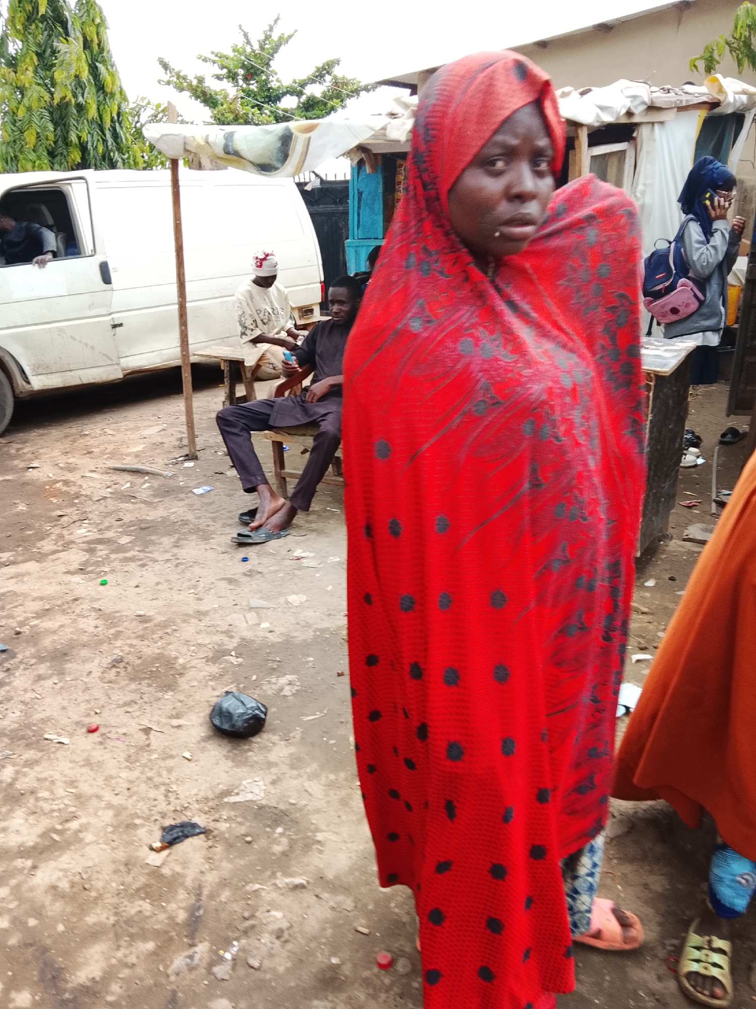 The image depicts a street scene, most likely in Abuja, Nigeria, given the style of dress of the people. A woman wearing a red headscarf with black polka dots is standing in the foreground. A man wearing a brown tunic and pants is seated on a wooden chair, barefoot with sandals nearby, under a makeshift shelter in the background. Another person wearing a white tunic and hat is sitting next to him on the same shelter. A white van is partially visible to the left of the men. Another person wearing a blue headscarf and pink backpack is partially visible on the right side of the frame. Debris and trash litter the ground.