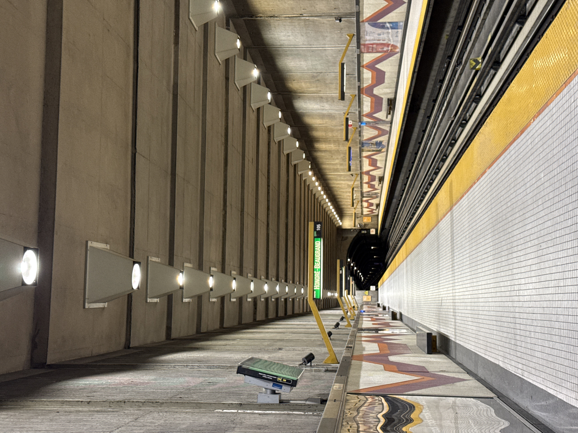 The image depicts the Honore-Beaugrand metro station platform in Montreal, Canada. The platform is empty. It shows a patterned floor along the platform, and a wall of white tiles opposite. The other wall is made of concrete with spaced lights. Station signage is also visible. Overhead are the train tracks.