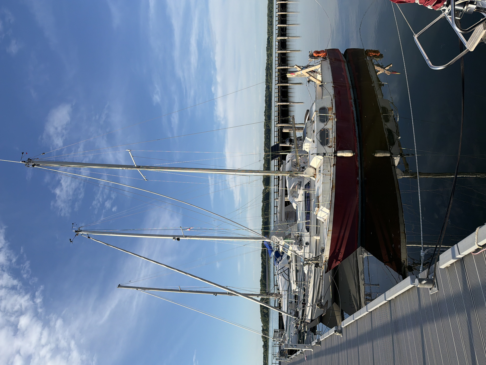 The image depicts a marina scene, likely in Ribnitz-Damgarten, Germany, during daylight. Several sailboats are docked side-by-side along a wooden pier. The sky is a clear blue with scattered, wispy white clouds, suggesting fair weather. The sun casts shadows on the pier, indicating it's either morning or late afternoon.
The foreground is dominated by the masts and rigging of the sailboats, which extend upwards towards the sky. The hulls of the boats are visible, with one prominently featuring a dark hull and another a lighter hull with a red stripe. Some boats have visible equipment like life preservers and solar panels. The water is dark and reflects the boats and the sky.
In the background, a line of pilings stretches into the distance along the water's edge, with a wooded shoreline beyond. The scene conveys a sense of tranquility and calm, typical of a marina setting. No people are visible in the image. There is no visible text in the image.