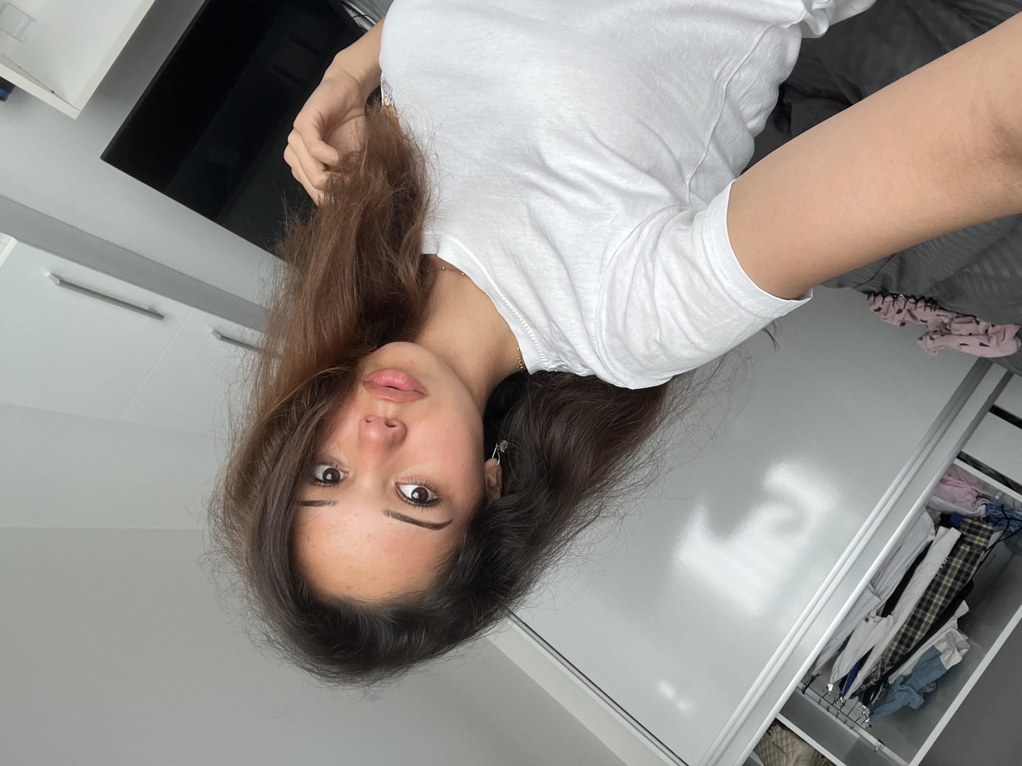 A young woman with long, dark brown hair and light brown eyes looks into the camera. She is wearing a white t-shirt and a silver necklace. She is in what appears to be a bedroom. A closet is visible to her left, filled with clothes. A bed with gray bedding is to her left. To her right, a black television screen is visible, mounted on a wall. The room is brightly lit, suggesting it is daytime. The overall scene is casual and personal. There is no information available to place this image in Zlatograd, Bulgaria.
