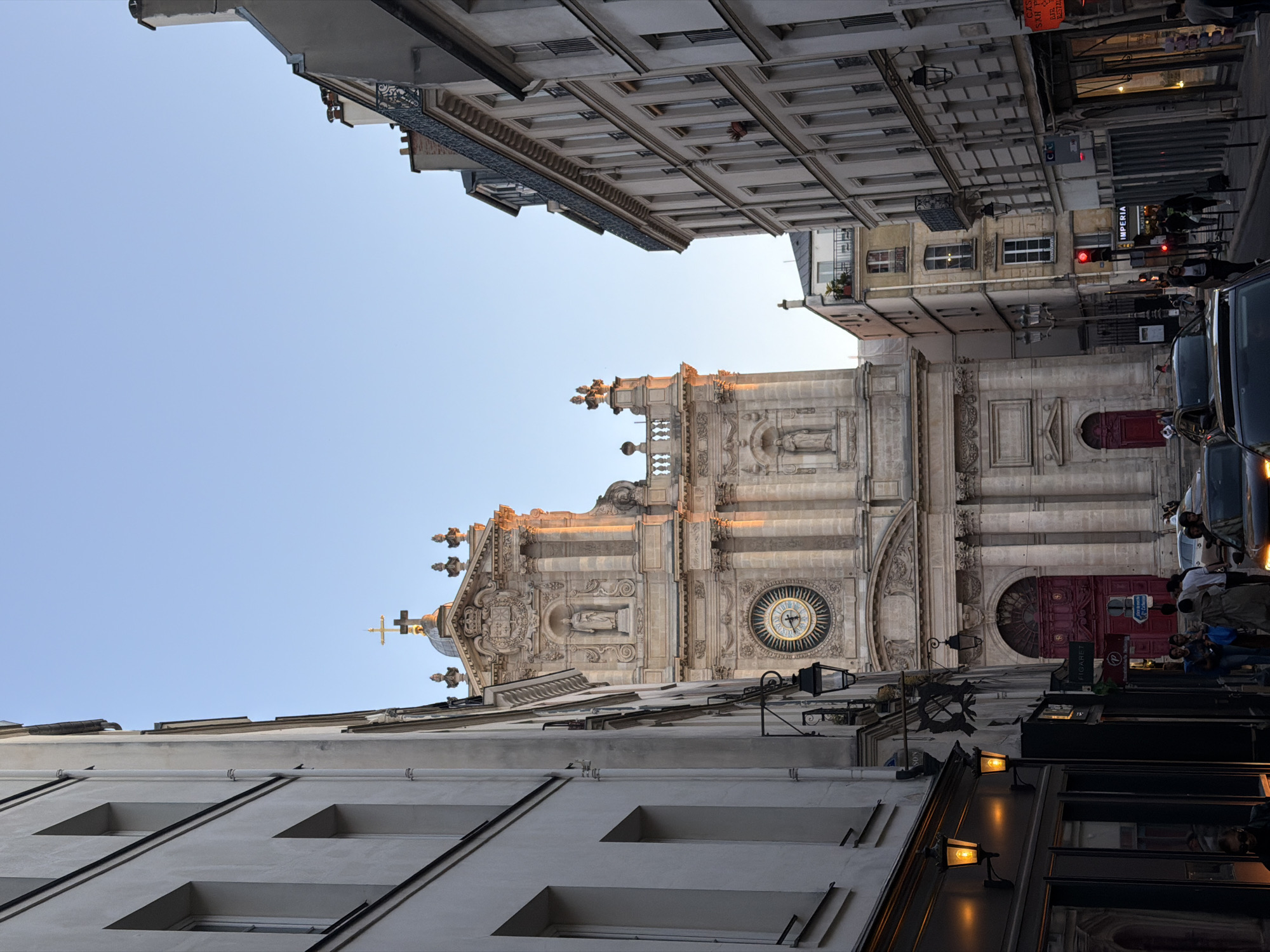 This outdoor photograph captures a bustling street scene in Paris, France, during what appears to be late afternoon or dusk, illuminated by a warm, golden light.
The central subject is a grand, ornately decorated church or cathedral, positioned on the right side of the frame. Its elaborate stone facade features intricate carvings, statues within niches, decorative pillars, balustrades, and a large circular clock face with Roman numerals. A golden cross tops a prominent dome or spire, catching the sun's rays.
Tall, multi-story Parisian buildings line both sides of the street. On the immediate left, a building with a simpler, light-colored facade features evenly spaced rectangular windows. Other buildings further down the street showcase classic Haussmannian architecture, complete with wrought-iron balconies and detailed cornices.
The street level depicts an active urban environment. Several cars are visible, both parked and potentially moving, including a dark vehicle on the right. Pedestrians can be seen walking along the sidewalks. A prominent red traffic light is visible on the right, and several streetlights, already illuminated with a warm glow, suggest the fading daylight.
Visible text includes "IMPERIA" on a storefront sign and "PIGALLE" on a distinctive red sign. Partially legible signs also show "Gros coq..." and "Restaurant". A notable red door is present near the base of the church or an adjacent building. The clear, light blue sky fills the upper-left portion of the image, complementing the warm tones of the architecture.