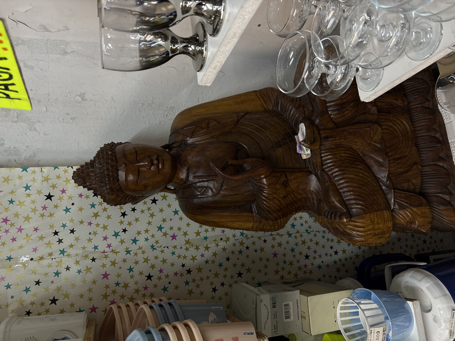 A large, carved, dark brown wooden Buddha statue, depicted seated with closed eyes and intricate robe details, is the central subject of a cluttered retail environment, likely in Buenos Aires, Argentina. The statue's left hand, resting in its lap, holds several folded Argentine Peso banknotes. To the left, stacked beige and blue plastic containers are visible above cardboard boxes, one bearing "INDUSTRIA ARGENTINA" and "Zero Waste" labels. A white sheet with a pattern of gold, pink, black, and teal stars serves as a backdrop. The right side features a textured, aged white wall with peeling paint. Two white shelves extend from this wall, displaying clear glassware: silver-stemmed goblets on the upper shelf and multiple clear coupe glasses on the lower. A yellow sign fragment with the green word "PAGA" is partially visible at the upper right.