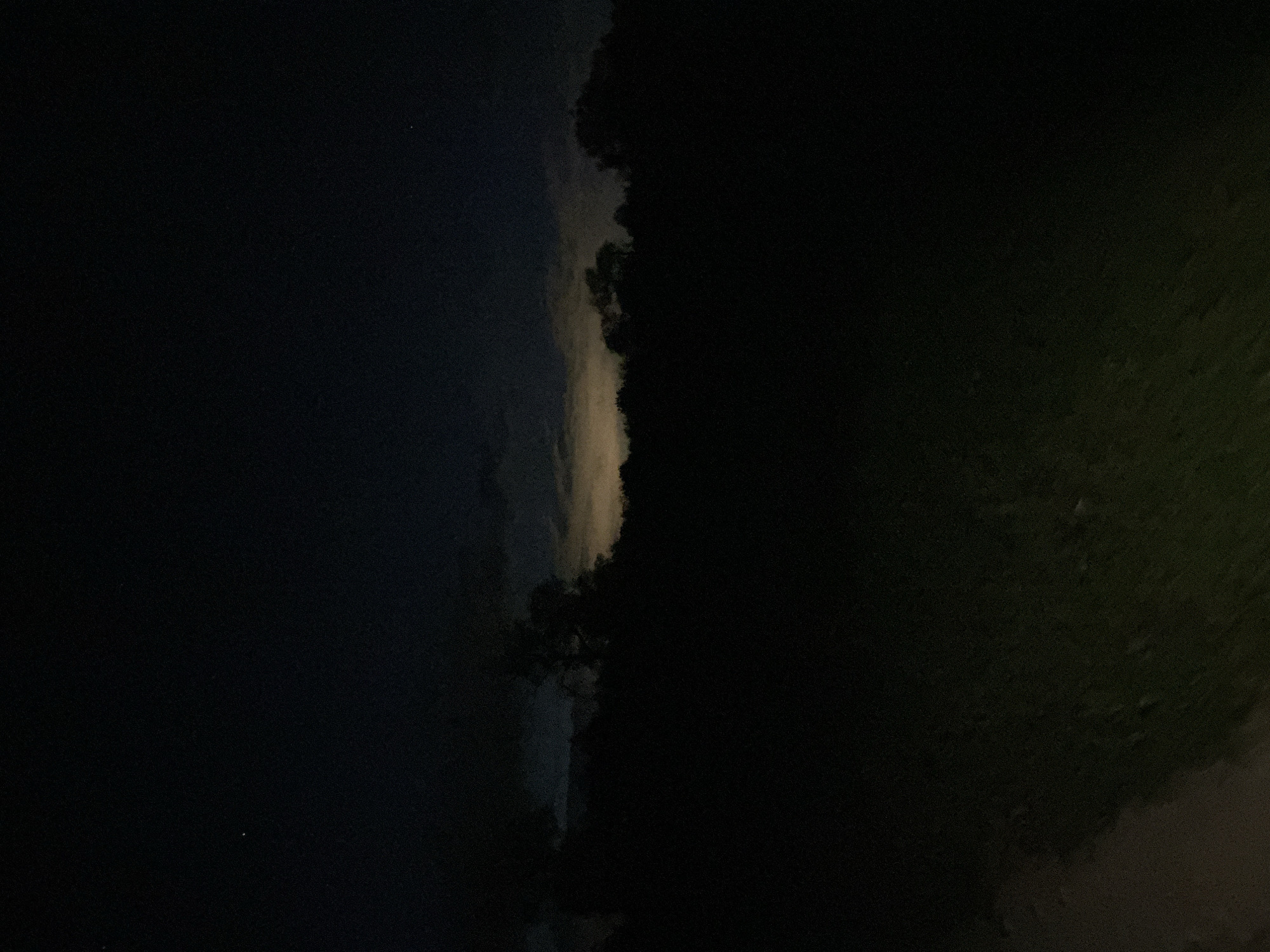 The image shows a dark scene, potentially taken at night in a natural environment, possibly Unknown, Unknown. There's a dark, cloud-filled sky on the left side of the image. The right side of the image is completely dark. In the center of the image, where the left and right zones meet, is what might be a border of tree-like shapes that seem to separate the sky from the ground area.