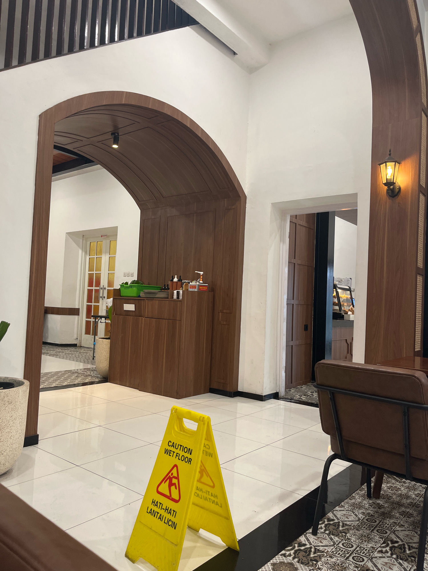 The image depicts the interior of a modern cafe with a warm, inviting ambiance. The architecture features a large arched wooden entryway, separating the main seating area from a walkway leading to a counter and a set of glass-paned doors. The flooring consists of white square tiles with black grout lines, and a patterned rug is visible in the foreground. A bright yellow "Caution Wet Floor" sign is prominently displayed in the center of the image, indicating potential hazard. To the right, a brown leather chair with a black metal frame is partially visible, suggesting a dining or seating area. The walls are painted white, contrasted by dark wood paneling and accents throughout the space. A vintage-style wall lamp with a warm yellow glow is mounted on the wood paneling. In the background, through an open doorway, a glimpse of a food display case can be seen. The scene appears to be clean and well-maintained, likely a public establishment.