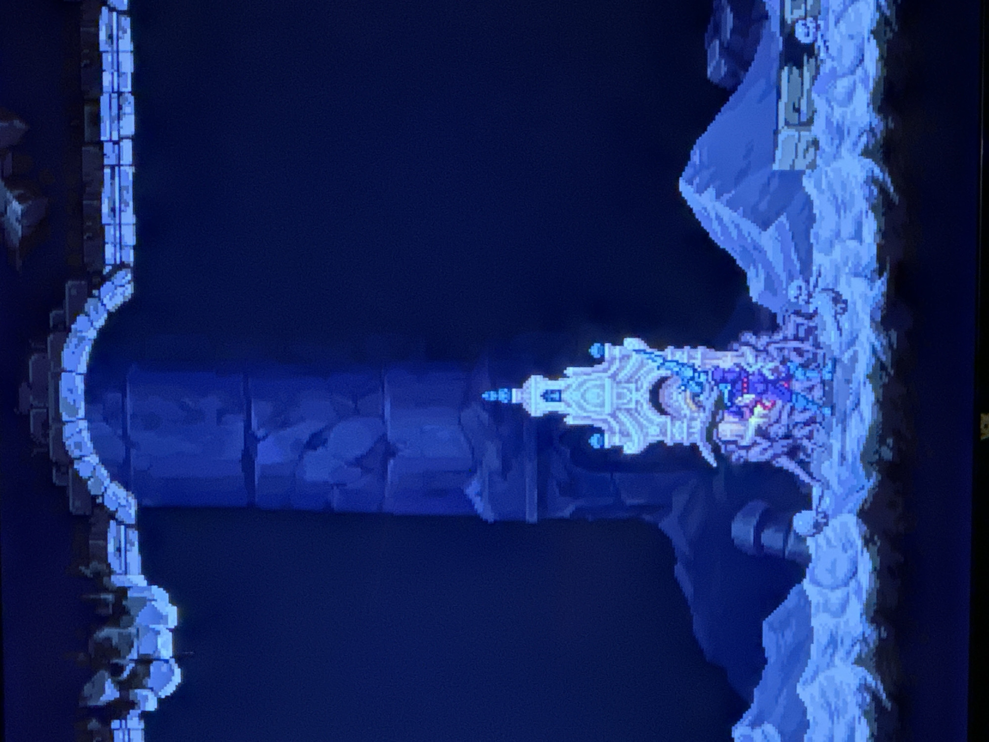 This image is a side-scrolling view of a video game environment featuring a blue-toned color palette. On the left, there is an arched, stone structure. A central, large, rectangular pillar extends horizontally from the left side towards the right. On the right, there is a structure appearing as if made of snow or ice, connected to the right side of the pillar. On top of this icy structure is a character or object with intricate, white and blue detailing, possibly with a smaller character atop it. The background is a dark blue, suggesting the game setting is underground or in a dimly lit area. The style appears to be pixel art. The image is not from the city of Novi Sad, Serbia.