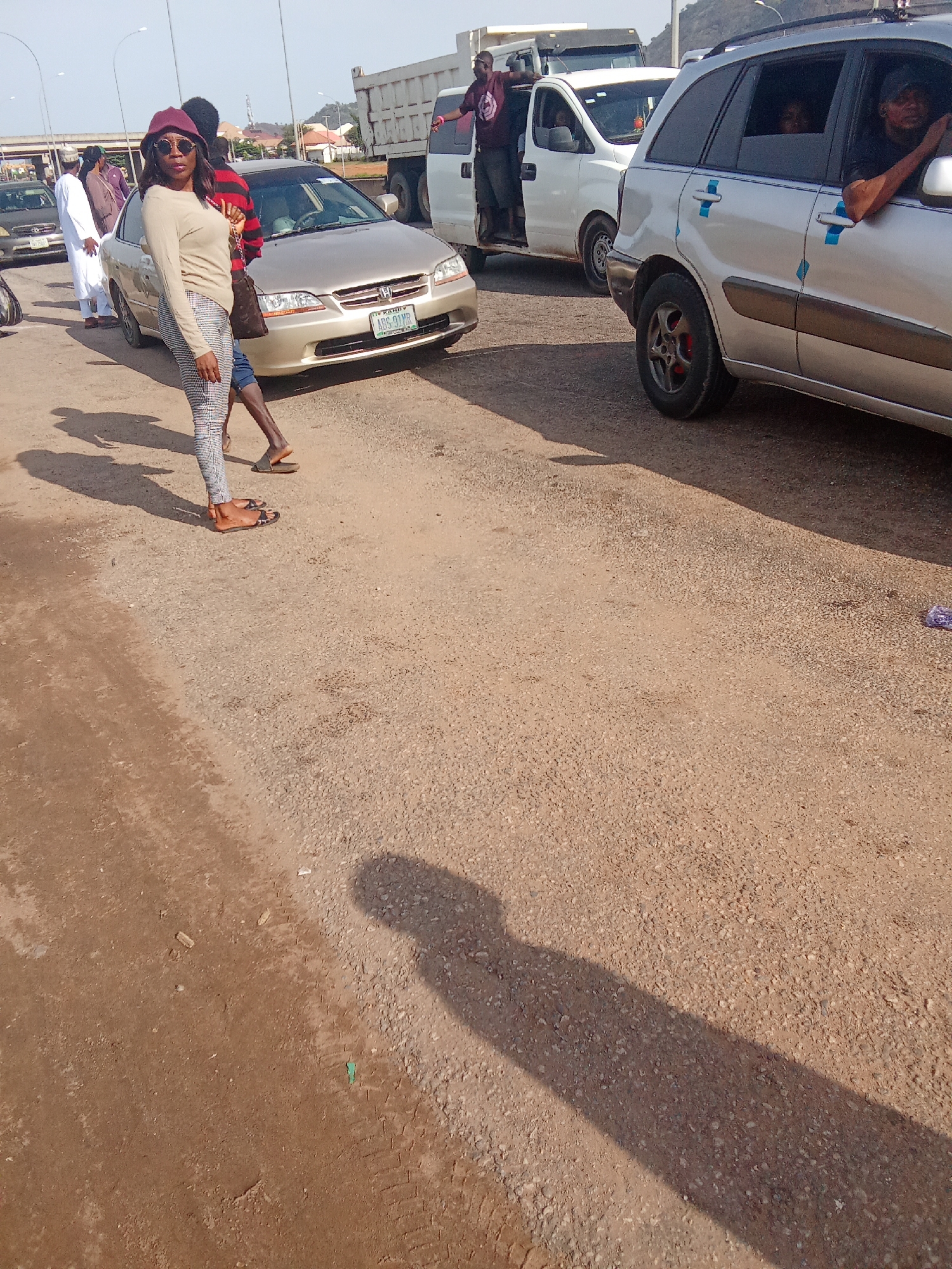 In Kagini, Nigeria, on a sunny day, a group of individuals are gathered around vehicles on a dusty road. A woman in a beige sweater, patterned leggings, sunglasses, and a burgundy hat stands near a gold sedan. Her right hand is on her hip, and her left hand is visible as she stands to the left of the vehicle. Behind her, a person in a red and black striped shirt is partially visible, and another individual in a white garment stands further back. A white truck and a silver SUV are also present. The SUV has blue tape on its side and a person with their arm out the window. The ground is covered in gravel and dirt. In the background, buildings and a hill are visible under a clear sky.