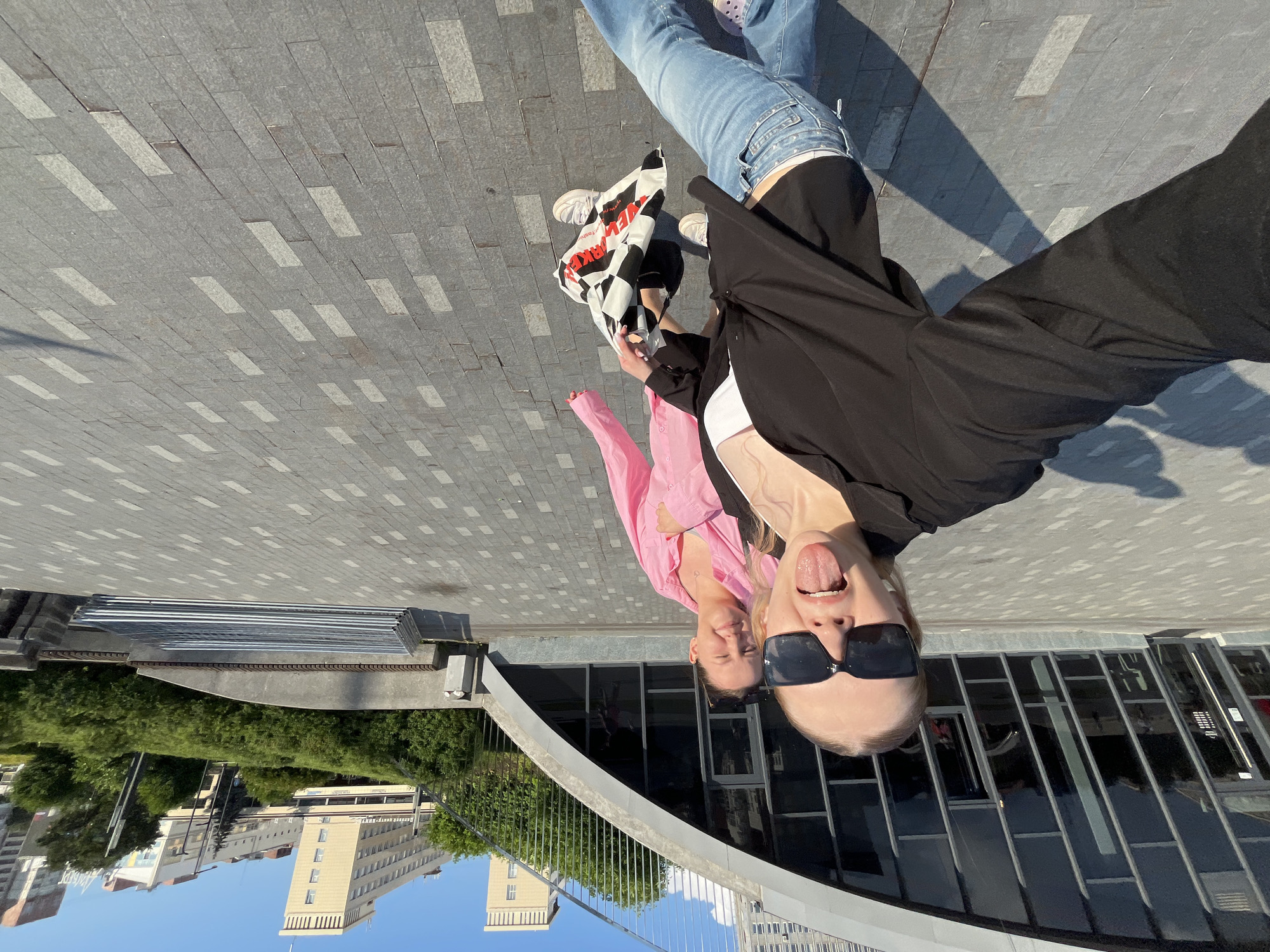 Two young women are standing on a wide paved area with a modern building with a curved roof and large glass windows to their left. The woman in the foreground is wearing a black open shirt, a white top, and blue jeans. She has large black sunglasses on and is sticking her tongue out at the camera. She is holding a black and white checkered bag. The woman behind her is wearing a bright pink shirt and has her hair pulled back. She is smiling. The background shows a tree-lined embankment with modern buildings beyond. This appears to be the embankment area of the Kama River in Perm, Russia, near the Perm State Art Gallery.