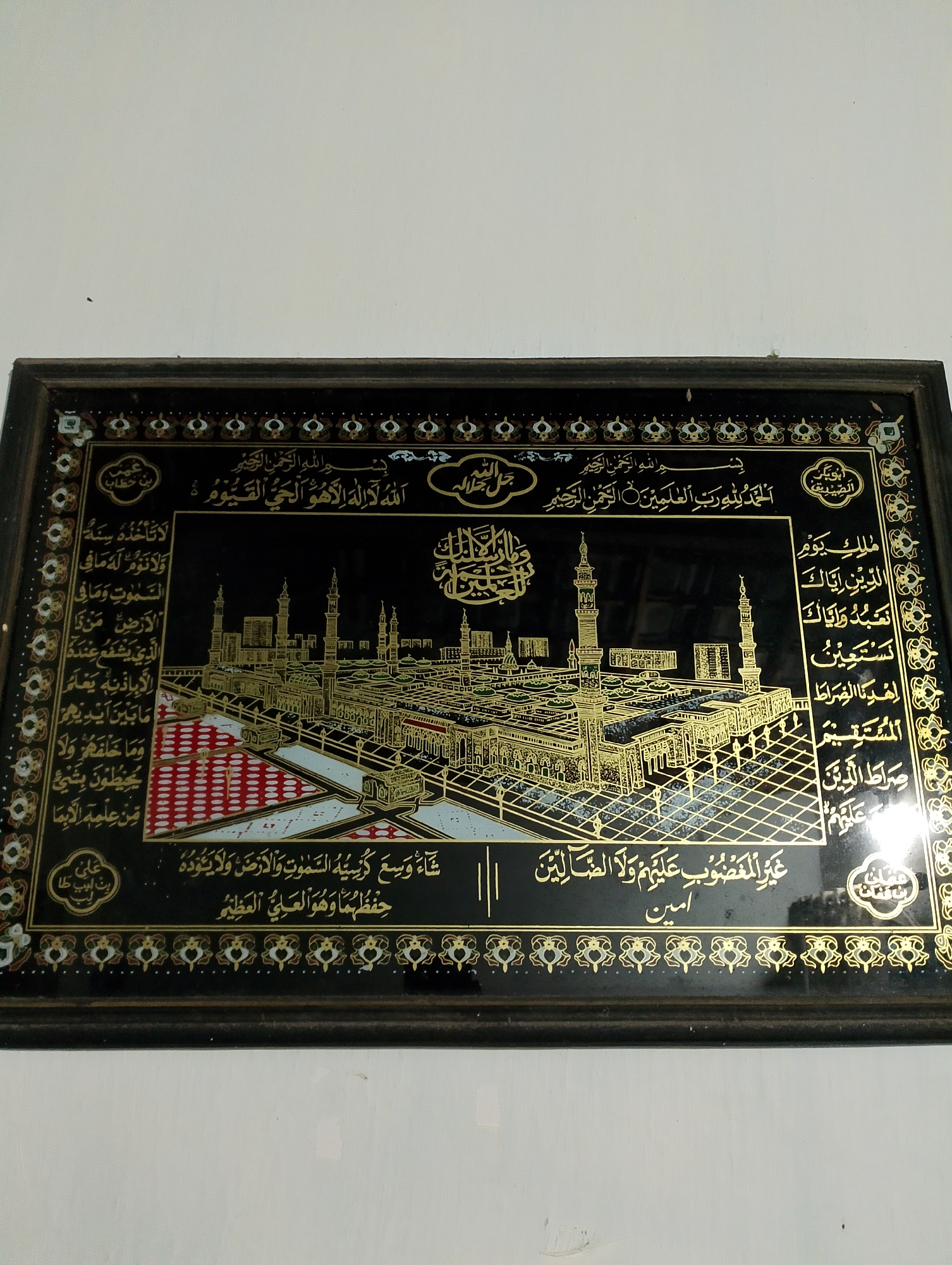 The image shows a framed artwork depicting a gold-embellished illustration of the Prophet's Mosque in Medina, Saudi Arabia, against a black background. The artwork is framed with a dark wood border and features Arabic calligraphy surrounding the illustration. On the left side of the frame, in a circular motif, are the words "عمر بن خطاب" (Umar bin Khattab), a prominent companion of the Prophet Muhammad. On the right side, also in a circular motif, are the words "ابوبكر الصديق" (Abu Bakr Al-Siddiq), the first Caliph. The artwork appears to be hanging on a plain white wall. The lighting suggests an indoor setting. The image itself is not from Rembang, Indonesia, but rather depicts a religious site significant in Islam.