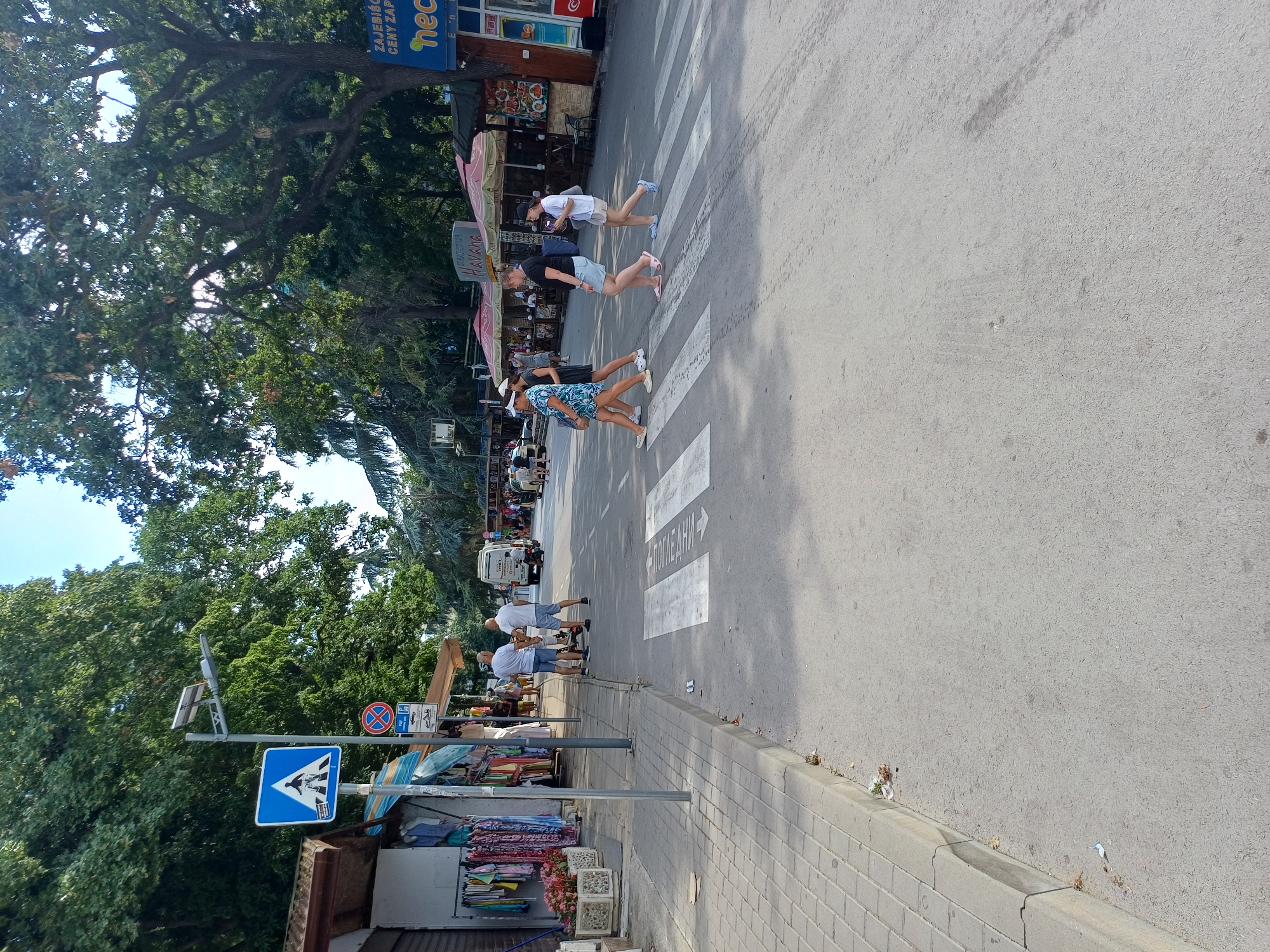 In Varna, Bulgaria, on a sunny day, people are crossing a street at a marked pedestrian crossing. The scene is a busy street with shops and trees lining one side. Several individuals are captured mid-stride, wearing casual summer attire. Some are walking in groups, while others are alone. Traffic signs are visible, including a blue sign with a pedestrian symbol. In the background, a van is stopped at the crossing, and other people are visible near shops. The overall atmosphere appears to be one of everyday activity in a coastal town. The text "НЕО" is visible on a sign in the background, likely indicating a business name. There are also some Russian words written on the street markings of the pedestrian crossing, which translates to "Look".