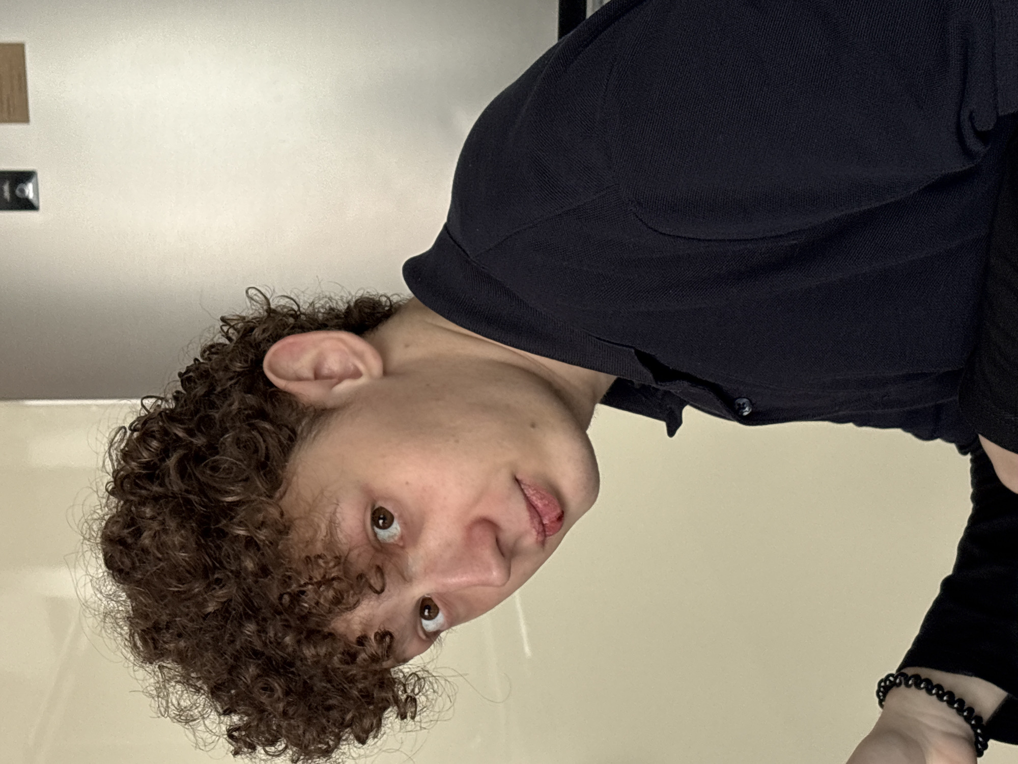 The image displays a head-and-shoulders portrait of a young individual, likely taken indoors in Ulyanovsk, Russia. The image is rotated 90 degrees clockwise. The subject has distinct, tightly curled medium-brown hair and light skin, with brown eyes looking directly at the viewer. They are wearing a dark, long-sleeved collared shirt, possibly black or dark blue, made of a textured fabric. A black beaded bracelet is visible on their right wrist. A notable detail is a chapped or slightly injured upper lip. The background consists of a plain, light-colored wall (off-white or light beige). In the upper left portion of the background, there are some indistinct shapes, including a small, dark rectangular object that appears to have faint, illegible markings, possibly "10|". The subject's expression is neutral, perhaps conveying curiosity or contemplation. There are no specific cues for time of day or weather, consistent with an indoor setting. No particular activity is taking place other than posing for the photograph.