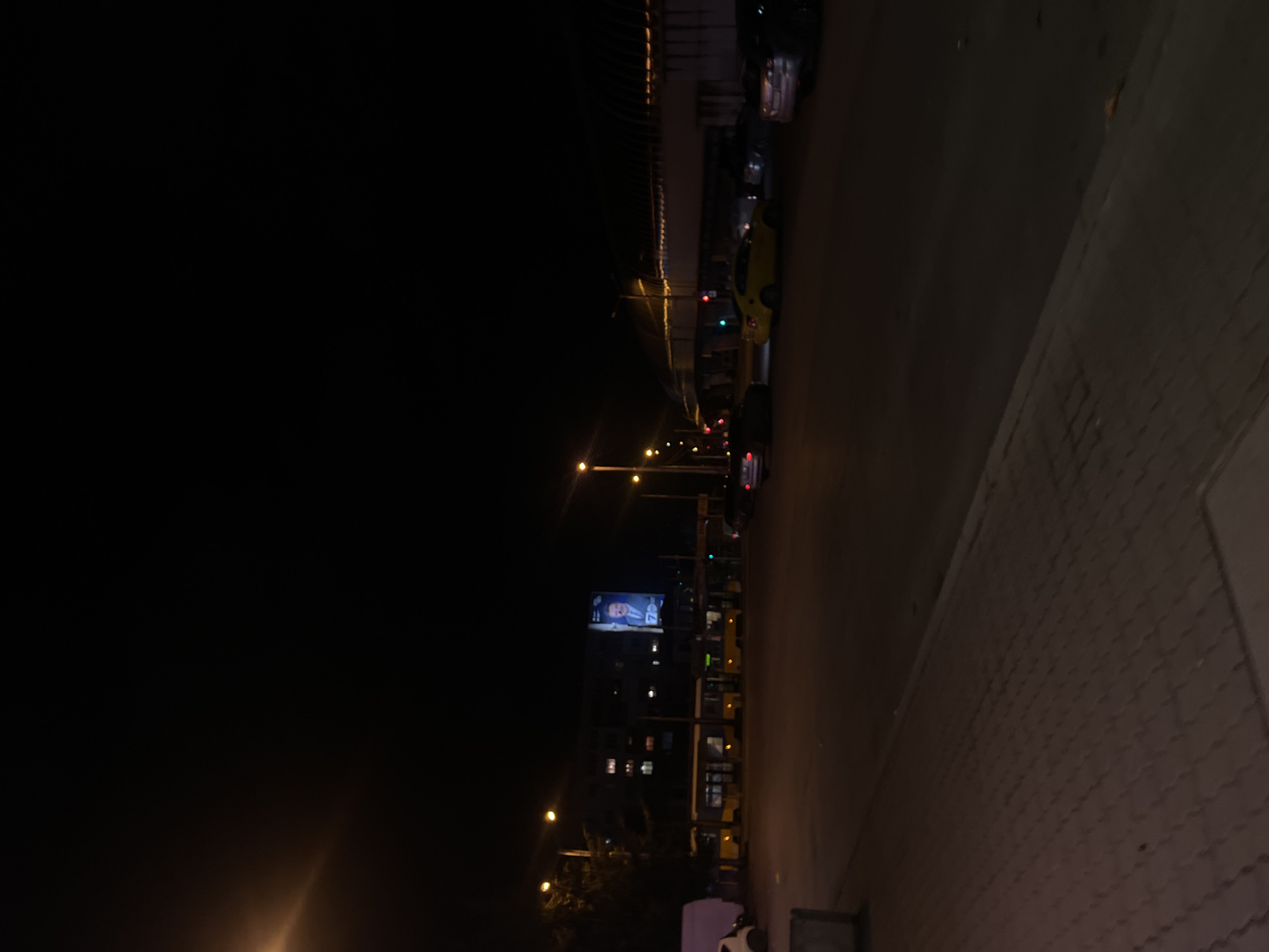 The image depicts a nighttime street scene in Sofia, Bulgaria. A line of parked cars and buses is visible along the right side of the road, adjacent to a paved sidewalk. Streetlights illuminate the road, and a large billboard displays an image, possibly a person's face. A multistory building with lit windows stands in the background. The sky is dark, with no visible stars.