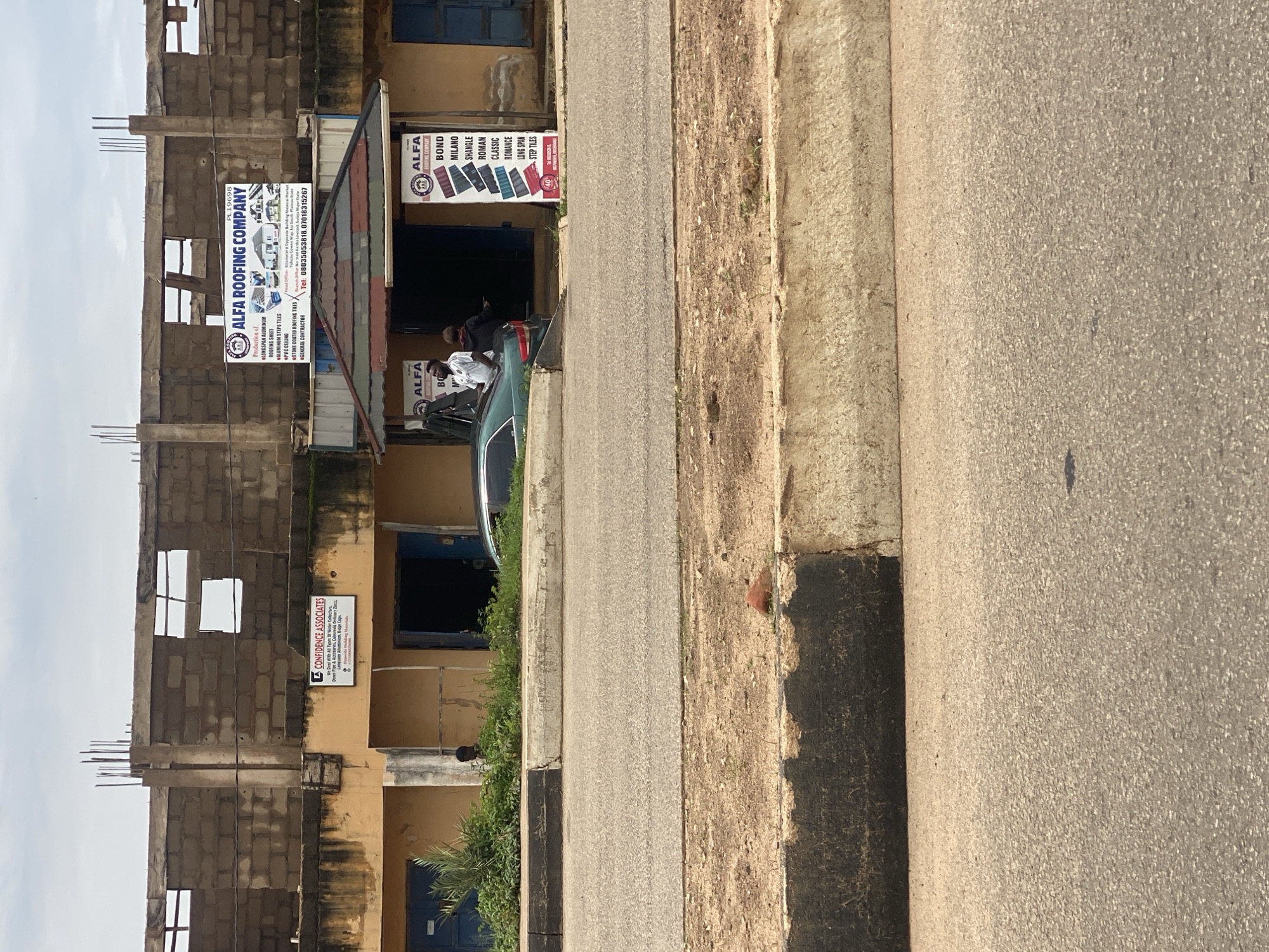 This image captures a street view in Gura Top, Nigeria, featuring a commercial building with prominent signage for "ALFA ROOFING COMPANY" and "CONFIDENCE ASSOCIATES." The building itself appears to be under construction or renovation, with exposed brickwork and visible rebar on the upper levels. The ground floor houses storefronts, with one entrance clearly marked by the Alfa Roofing Company's banner, which lists various roofing materials and services including "ROOFING SHEET," "ALUMINUM STEPS TILES," "PVC CEILING," "STONE COATED ROOFING TILES," and "GENERAL CONTRACTOR."
Another sign for "CONFIDENCE ASSOCIATES" advertises "All Types Of Water Collectors," "Down Pipe & Accesaries," and other related building materials. Next to the main building, a second Alfa Roofing Company sign displays different tile styles like "BOND," "MILANO," "SHANGLE," "ROMAN," "CLASSIC," "ROMANCE," "LONG SPAN," and "STEP TILES."
In front of the building, a car is parked near the entrance. A few individuals are visible near the car and the storefronts, suggesting business activity. The scene is illuminated by daylight, and the weather appears to be overcast or cloudy. The presence of the road, sidewalk, and building indicates an urban or semi-urban setting. The overall impression is one of a commercial hub for construction and building materials in Nigeria.