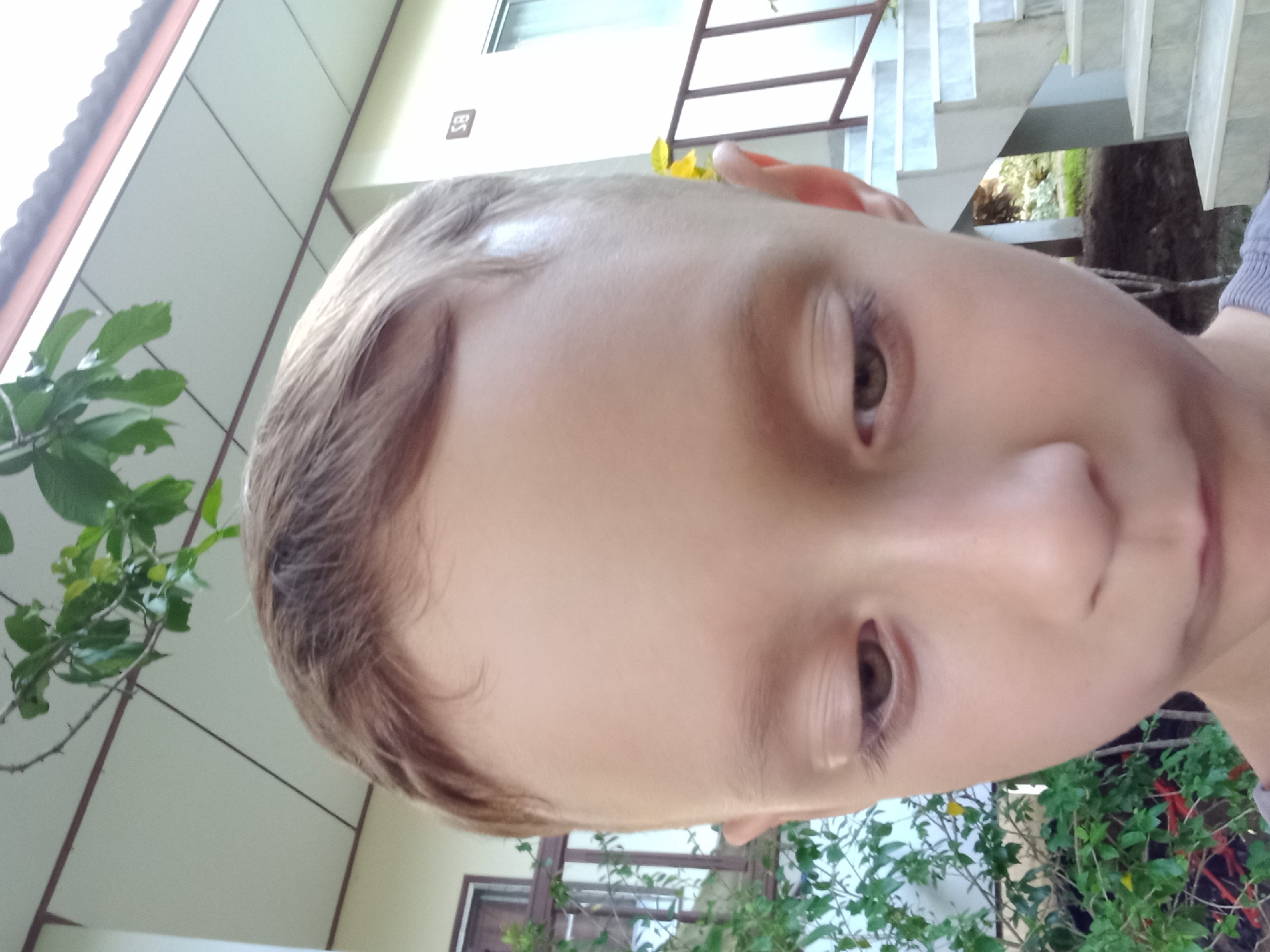 An extreme close-up, eye-level shot focuses on the face of a young boy. His head is tilted downward, and his gaze is directed away from the camera, obscuring his eyes. The boy has light brown hair and fair skin. Behind him, to the left, is a building with a cream-colored facade and a sloped roof. A railing with brown wooden posts and railings is visible, along with lush green foliage. To the right, a white staircase with grey marble steps leads upwards. A number "58" is affixed to the building's wall. The overall setting suggests an outdoor environment, possibly residential, in Baan Mae Nam, Thailand.