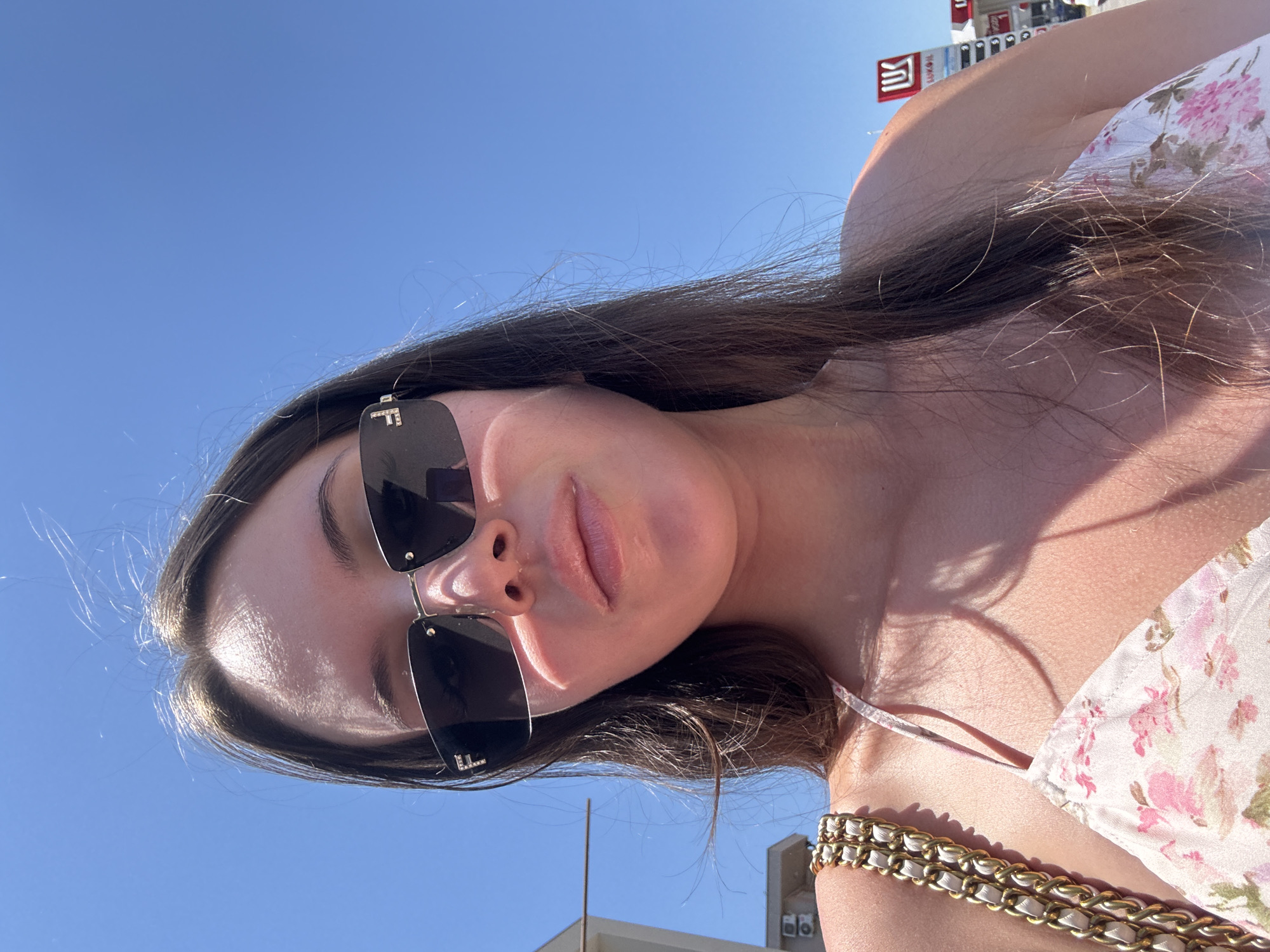 A close-up, low-angle outdoor selfie shows a young woman from the chest up, against a bright blue sky. She is wearing dark, rectangular sunglasses with the letter "F" on them and a floral print dress with pink and white flowers. A gold chain strap purse is visible on her left shoulder. Sunlight casts shadows across her skin and hair, which is long and brown. In the background, a building with air conditioning units is visible to the left, and a Lukoil gas station sign is to the right. The overall scene suggests a warm, sunny day in an urban environment.