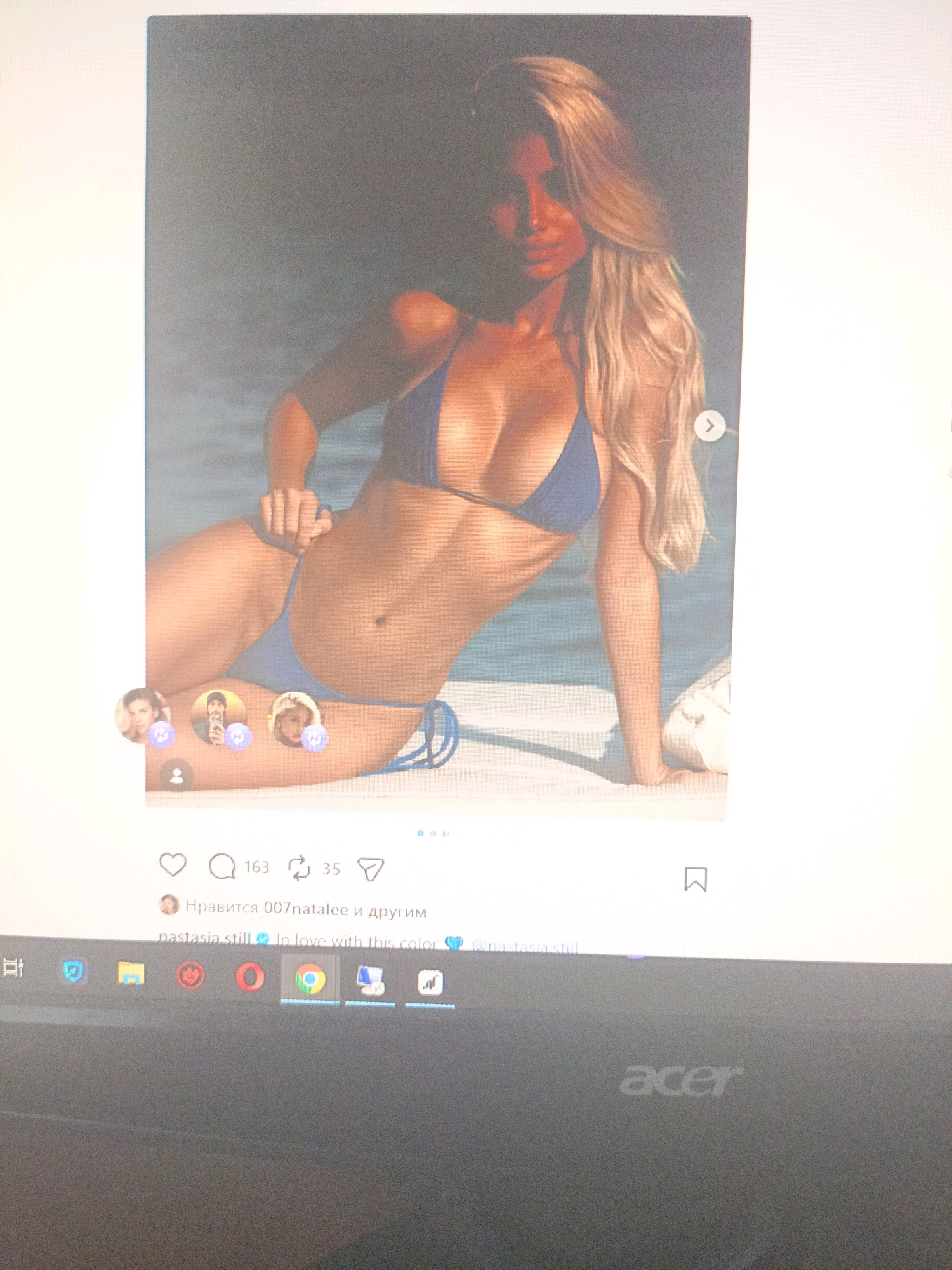 The image shows a screenshot of an Instagram post on a laptop screen. The post features a woman in a blue bikini posing on what appears to be a beach or poolside. The woman has long blonde hair and is looking towards the camera. Below the image, there are social media icons for likes, comments, and shares. A caption reads "nastasia.still In love with this color." The lower part of the laptop screen shows a taskbar with various application icons, including Google Chrome. There is no information in the image to suggest the location is Ухманы, Russia.